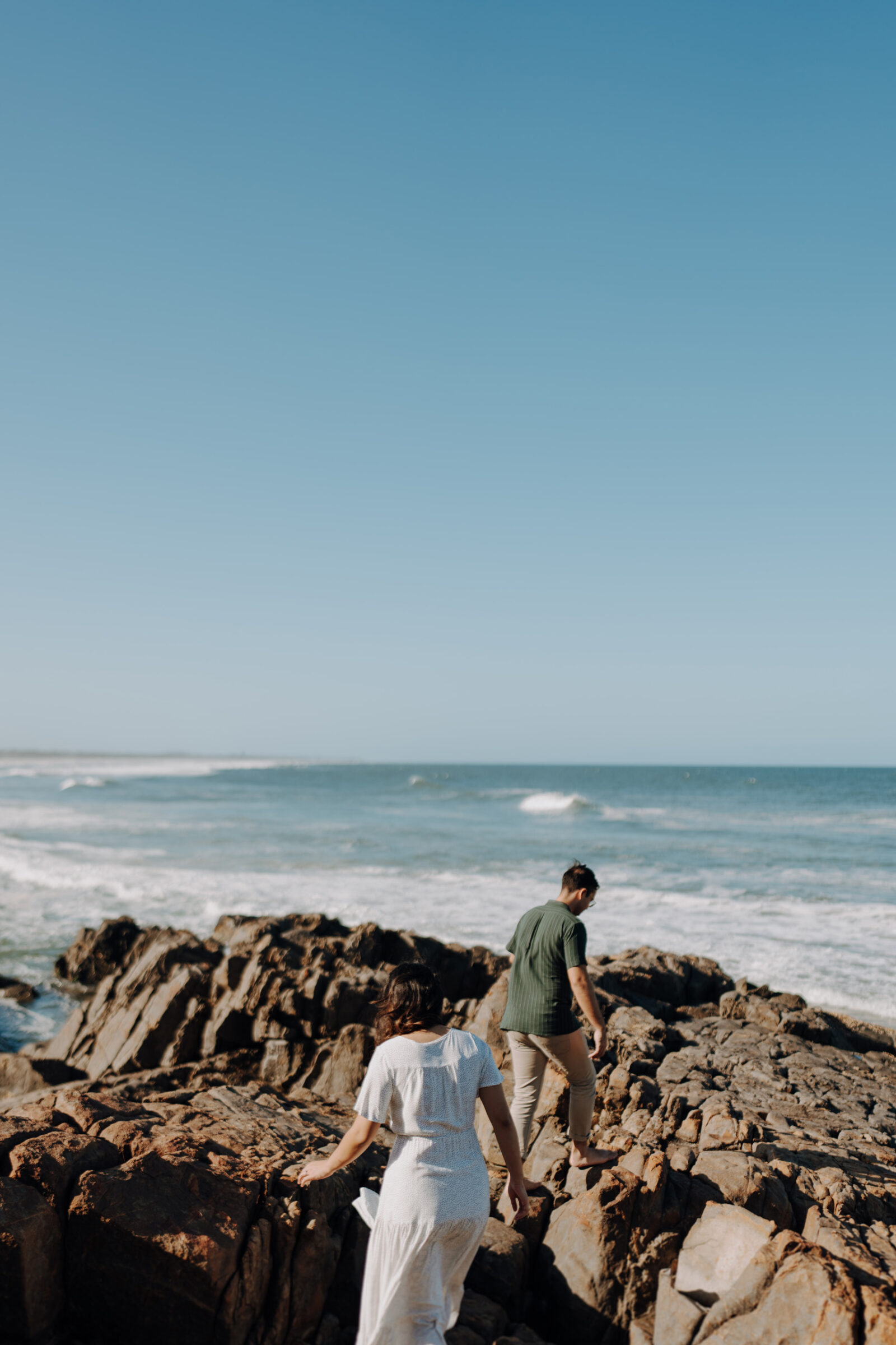 Two people walking on rocky shoreline with ocean waves in the background under a clear blue sky.