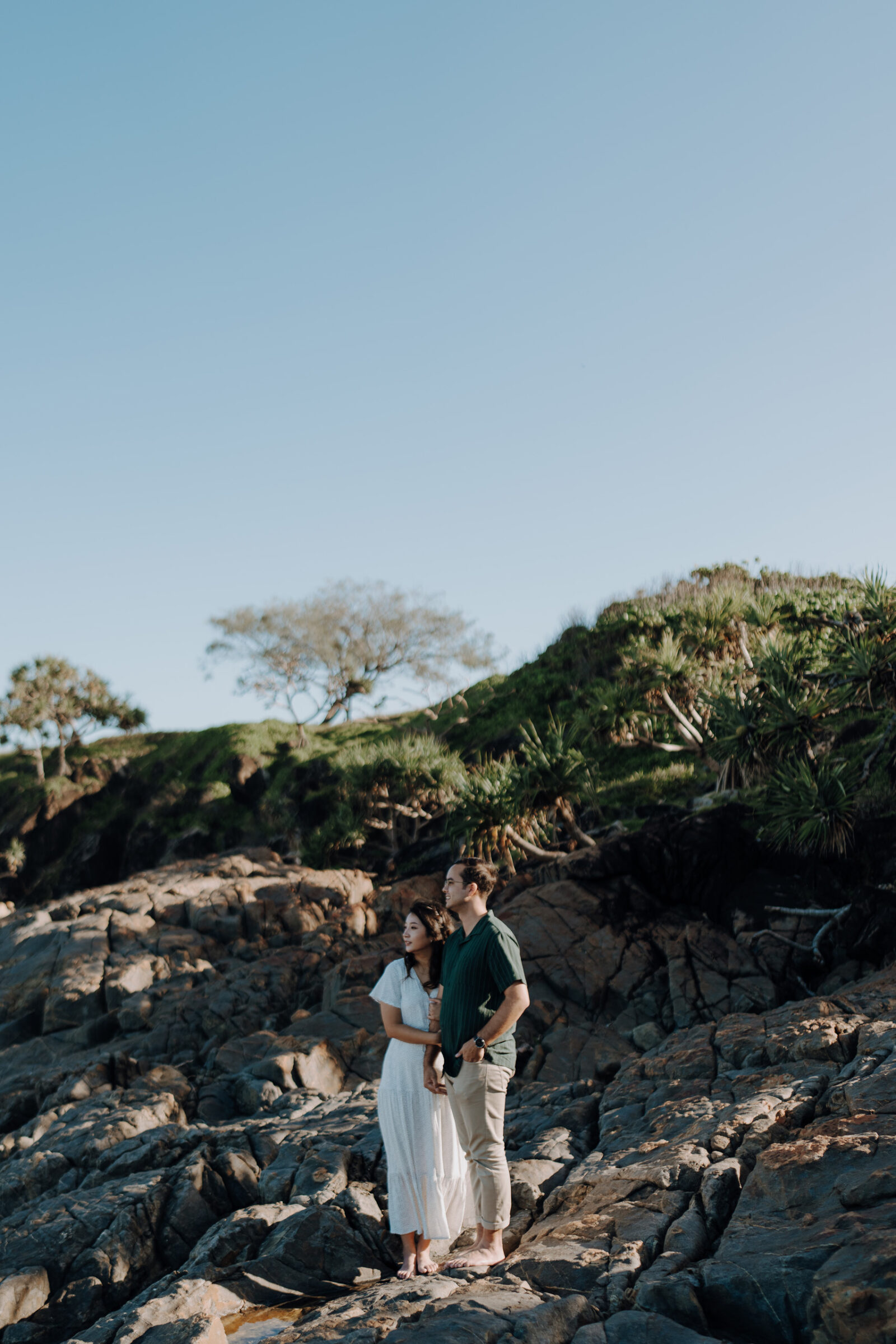 A couple stands on rocky terrain, with greenery and trees in the background under a clear blue sky.