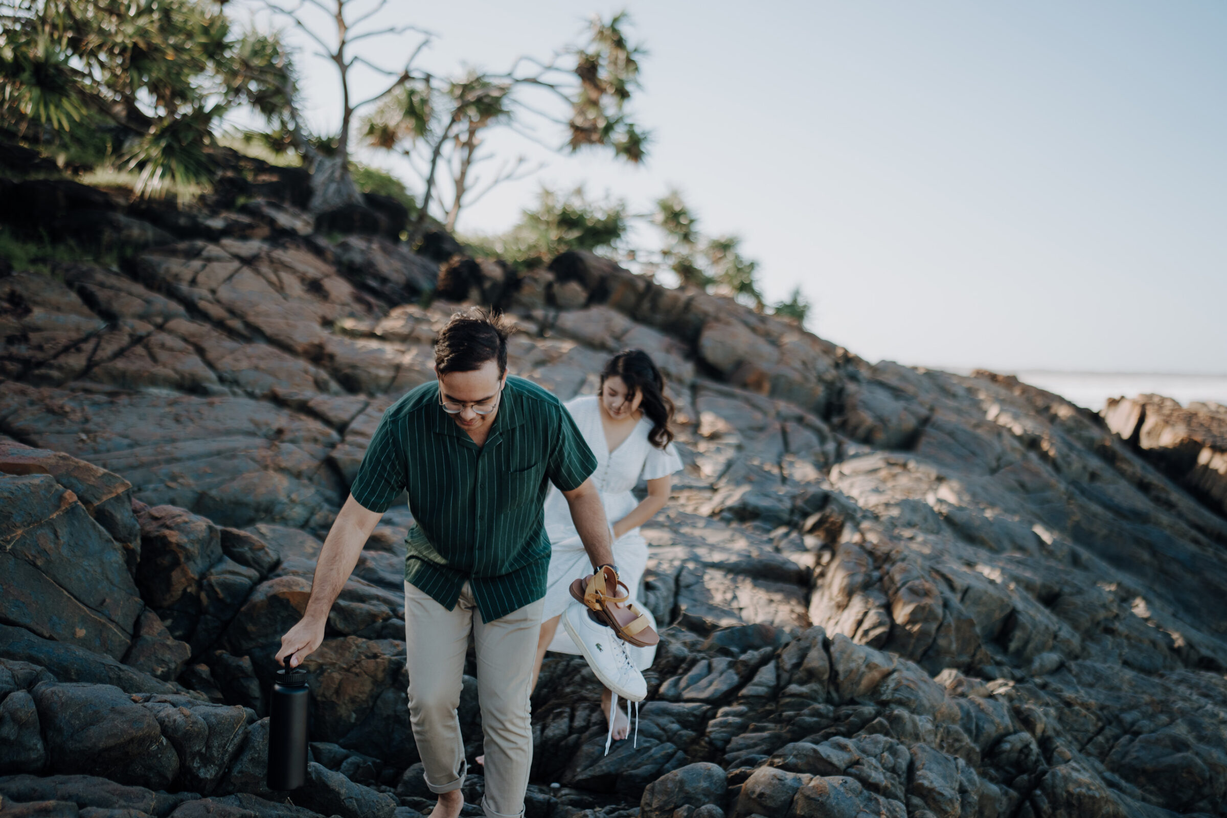 A man and woman walk barefoot over rocky terrain. The man carries a drink container and a pair of shoes. Sparse trees are visible in the background.