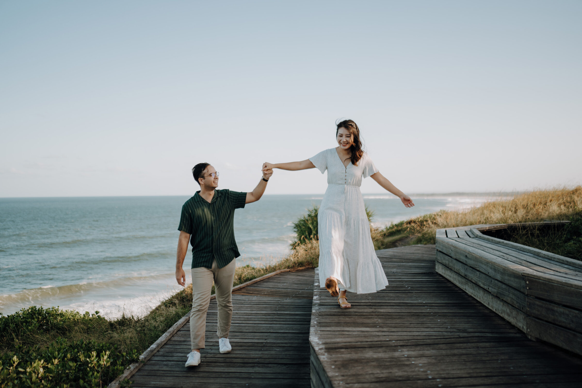 A couple walks hand in hand along a wooden boardwalk by the ocean, with the woman balancing on the edge.