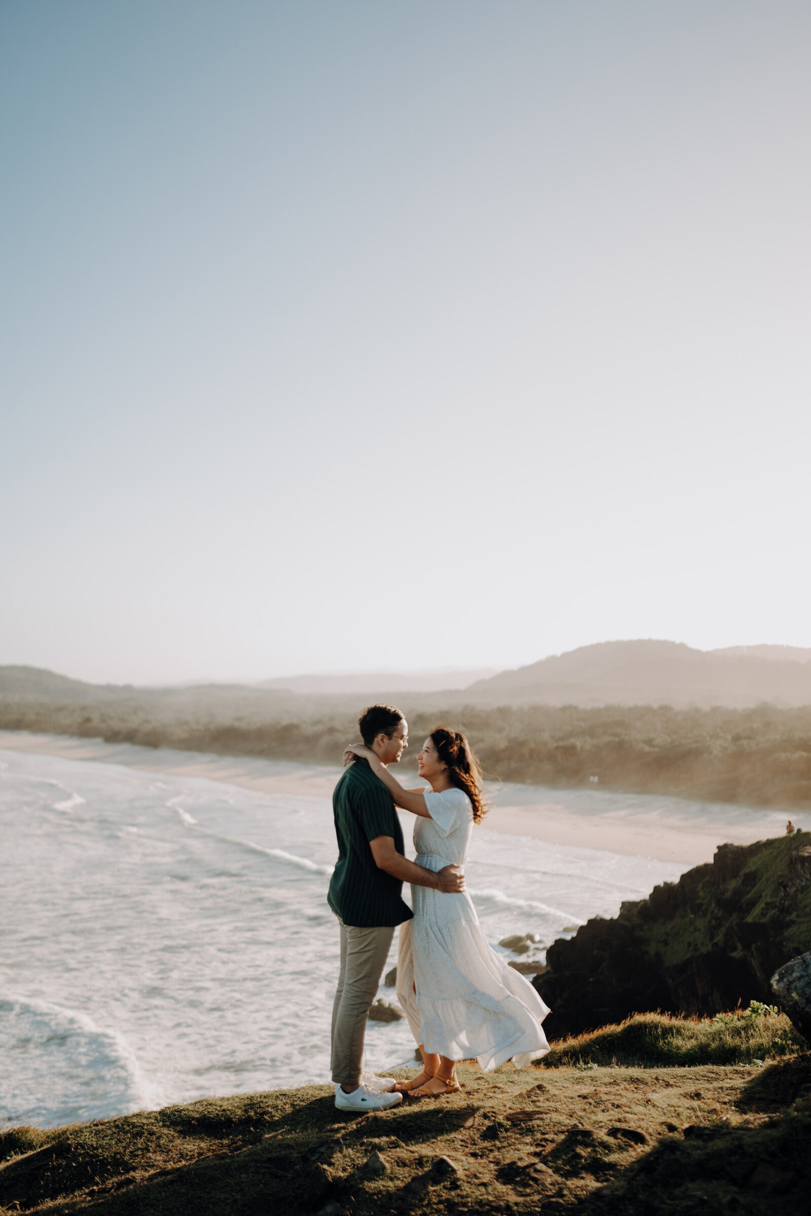 A couple stands embracing on a cliff overlooking the ocean, with a sandy beach and mountains in the background.