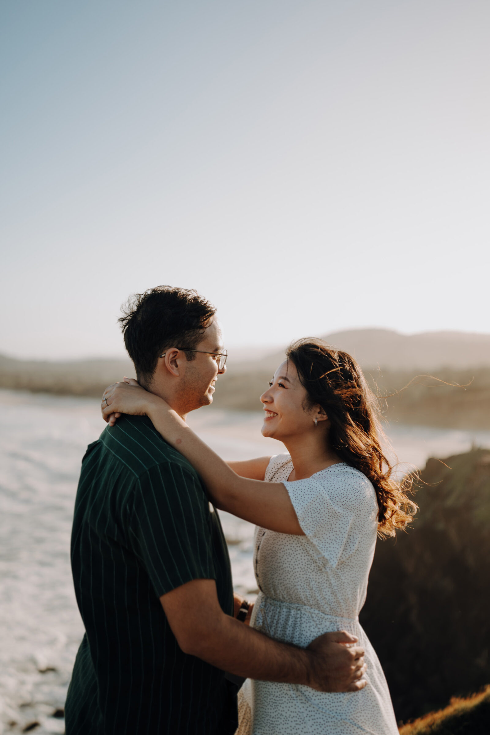 A couple embraces by the seaside, looking at each other and smiling, with the ocean and distant hills in the background.