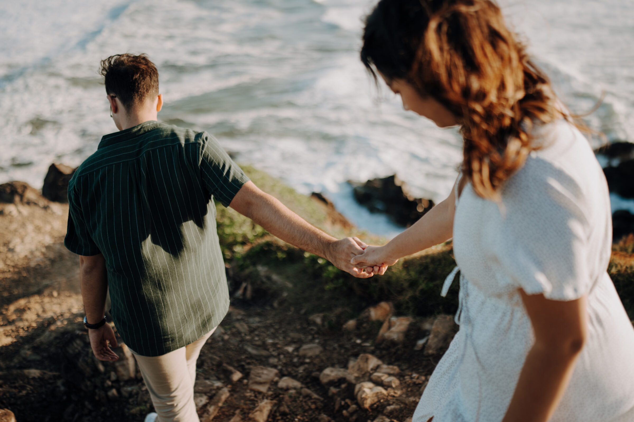 A couple walks hand in hand down a rocky path near the ocean.