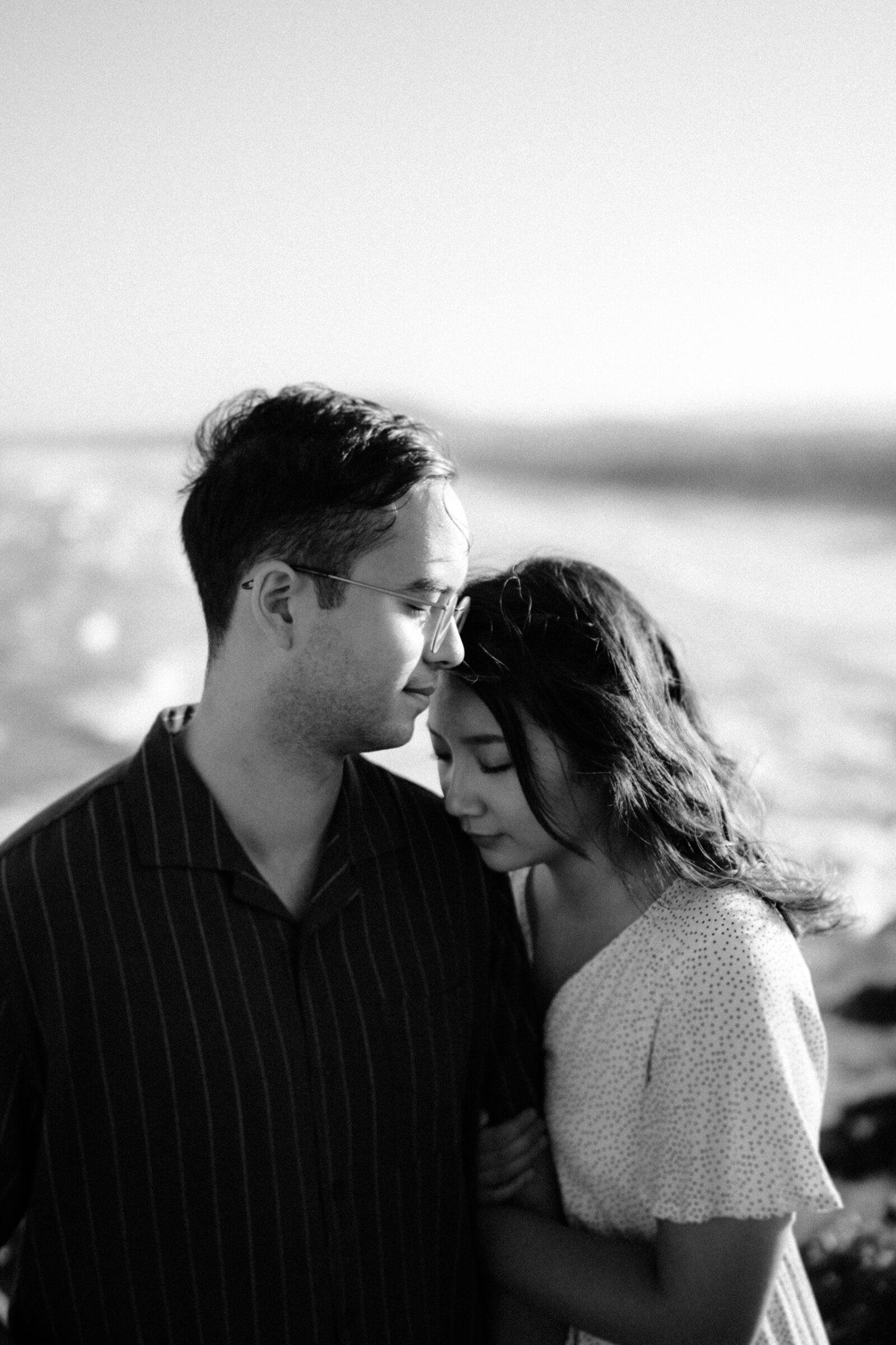 A couple stands closely together on a beach, with the woman resting her head on the man's shoulder. The scene is in black and white.