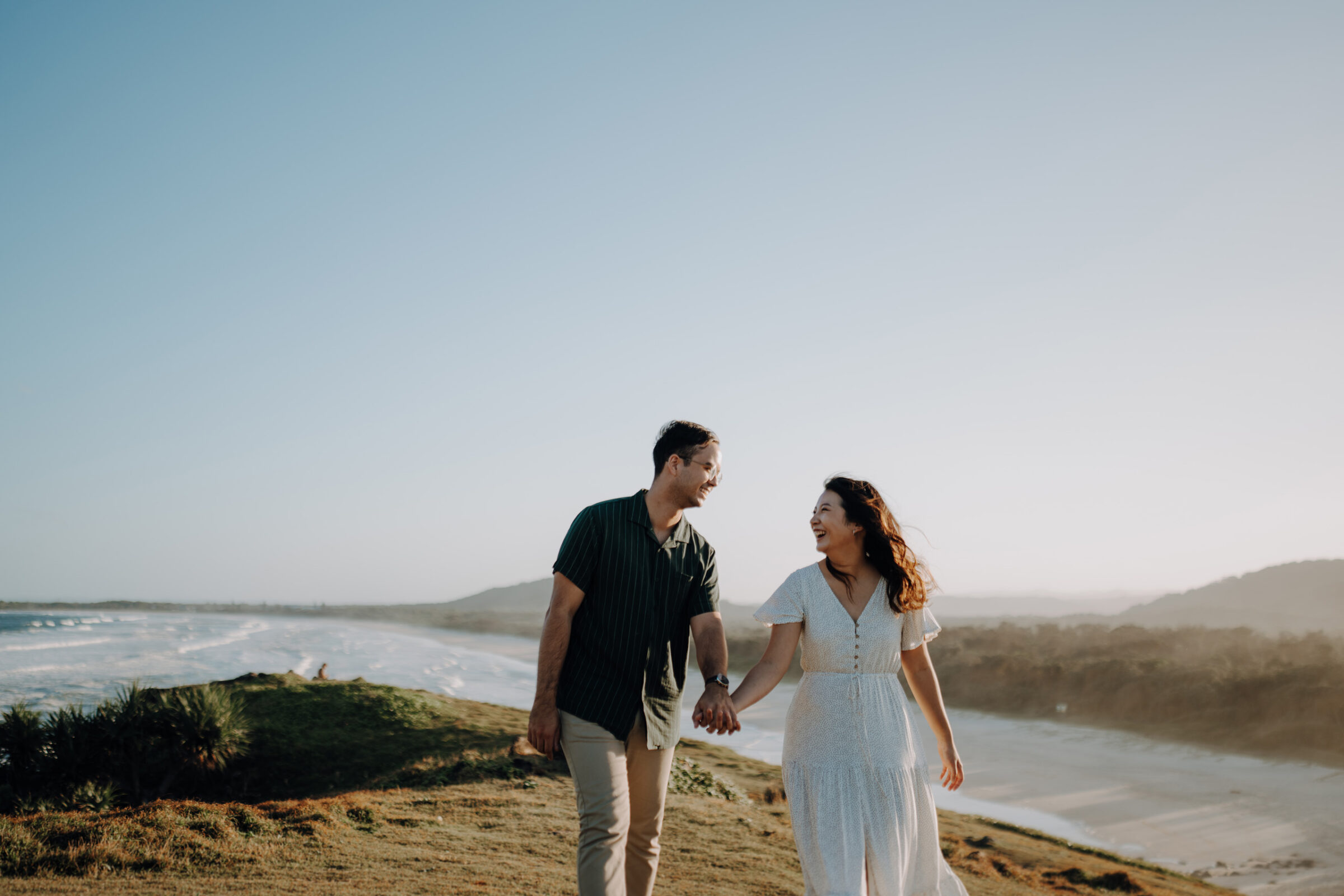 A couple holds hands, walking on a grassy hill by the ocean, with a clear sky and waves in the background.