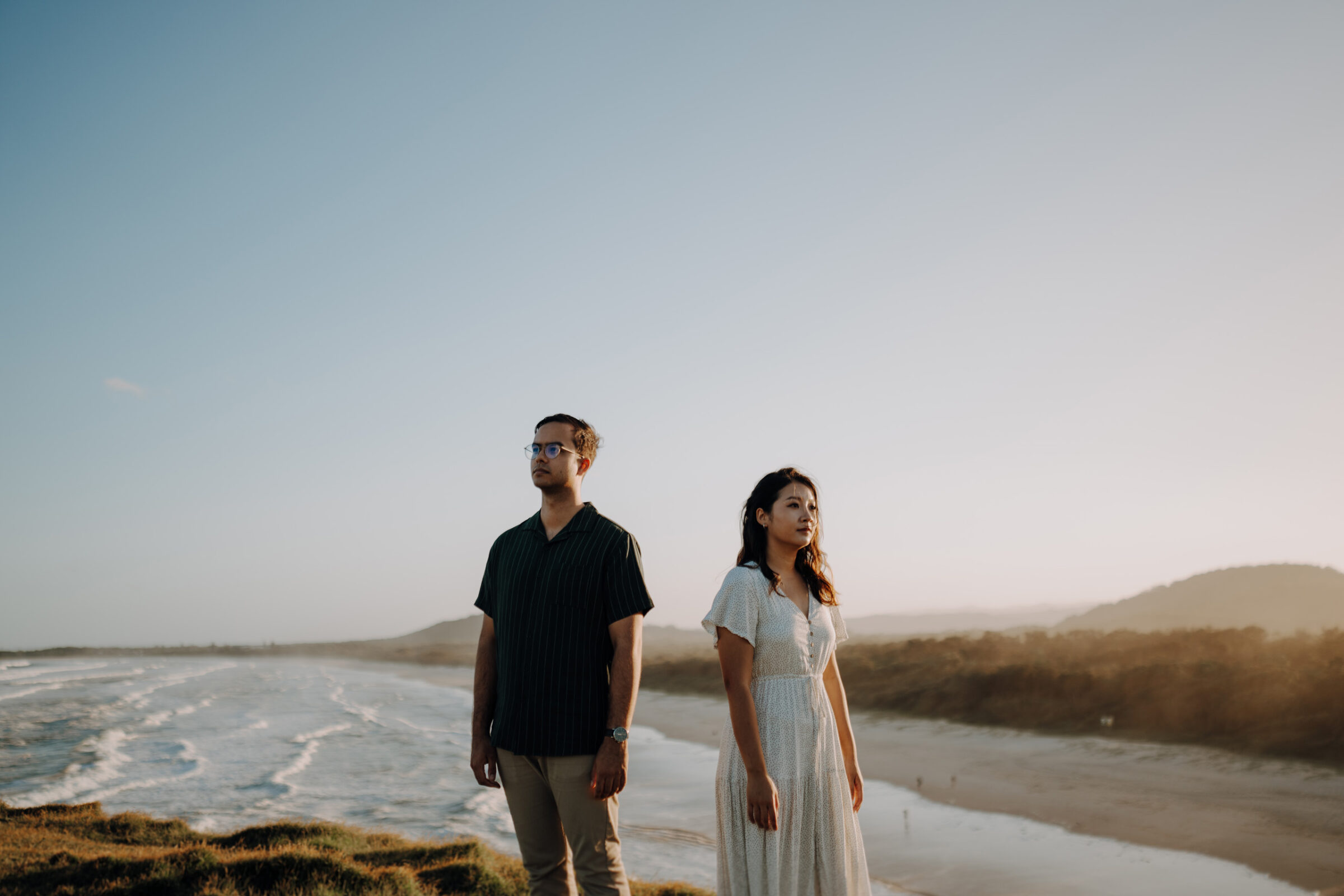 Two people stand on a cliff overlooking the ocean, with a clear sky and distant hills in the background.