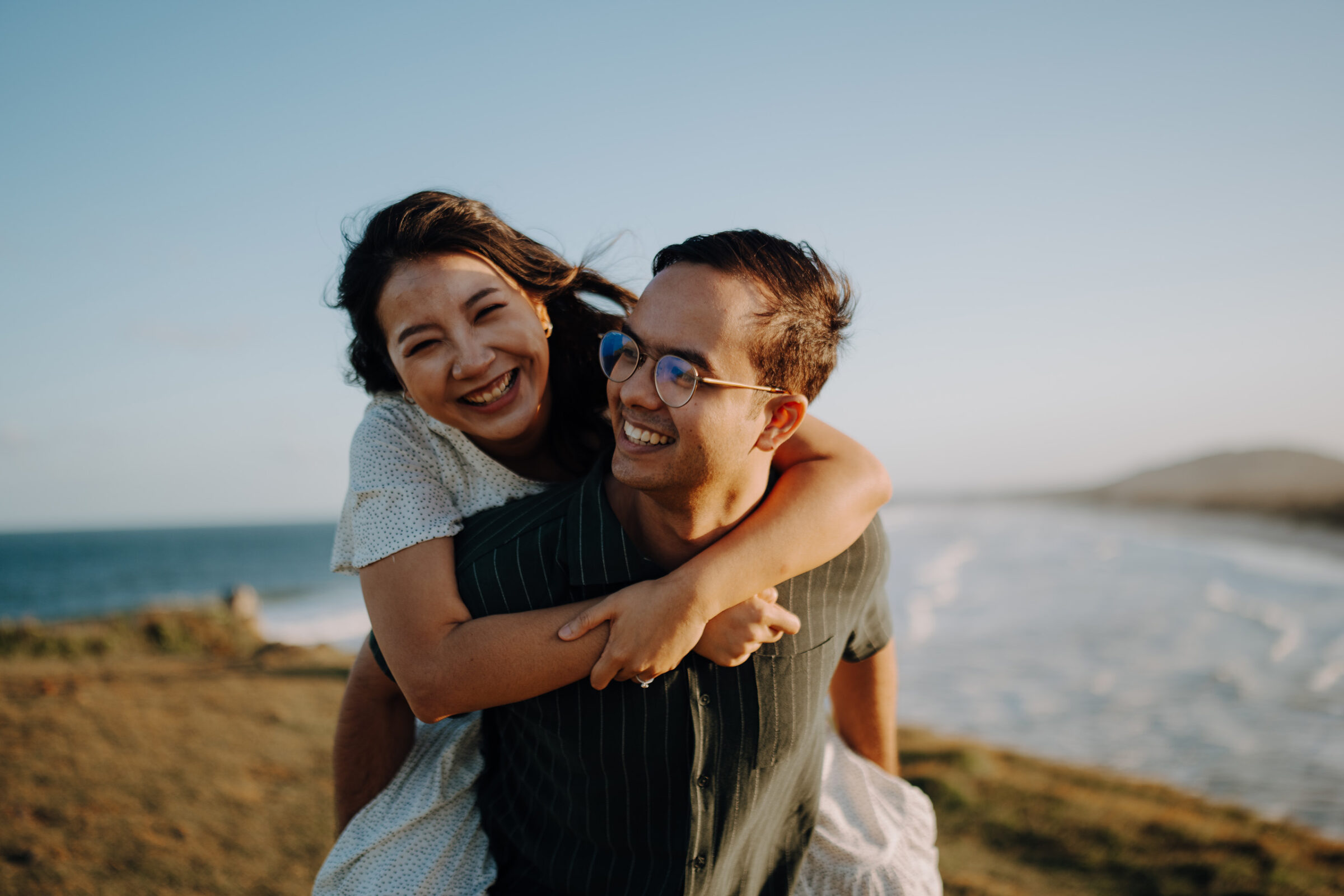 A woman playfully piggybacks on a man near a coastline. Both are smiling, with the ocean and sky in the background.