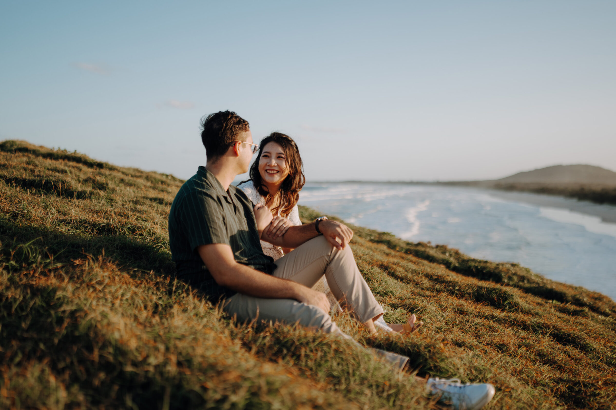 A couple sits on a grassy hillside by the beach, looking at each other and smiling, with the ocean in the background under a clear sky.