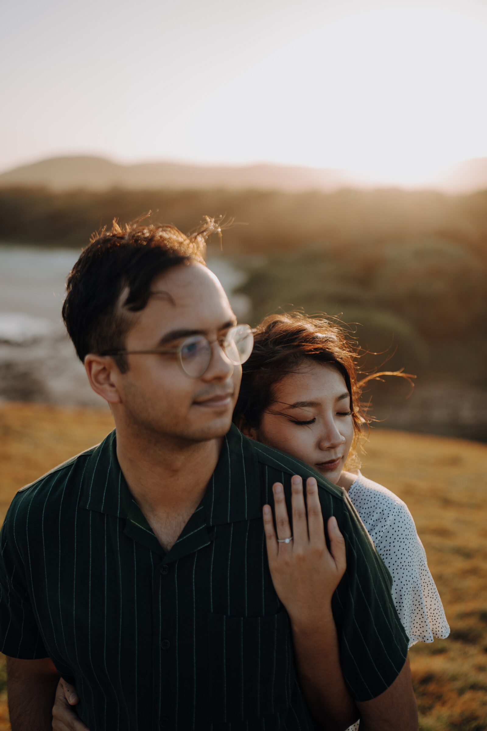 A couple stands in a field at sunset; one person, wearing glasses and a green shirt, faces forward while the other, in a white dress, gently embraces them from behind.