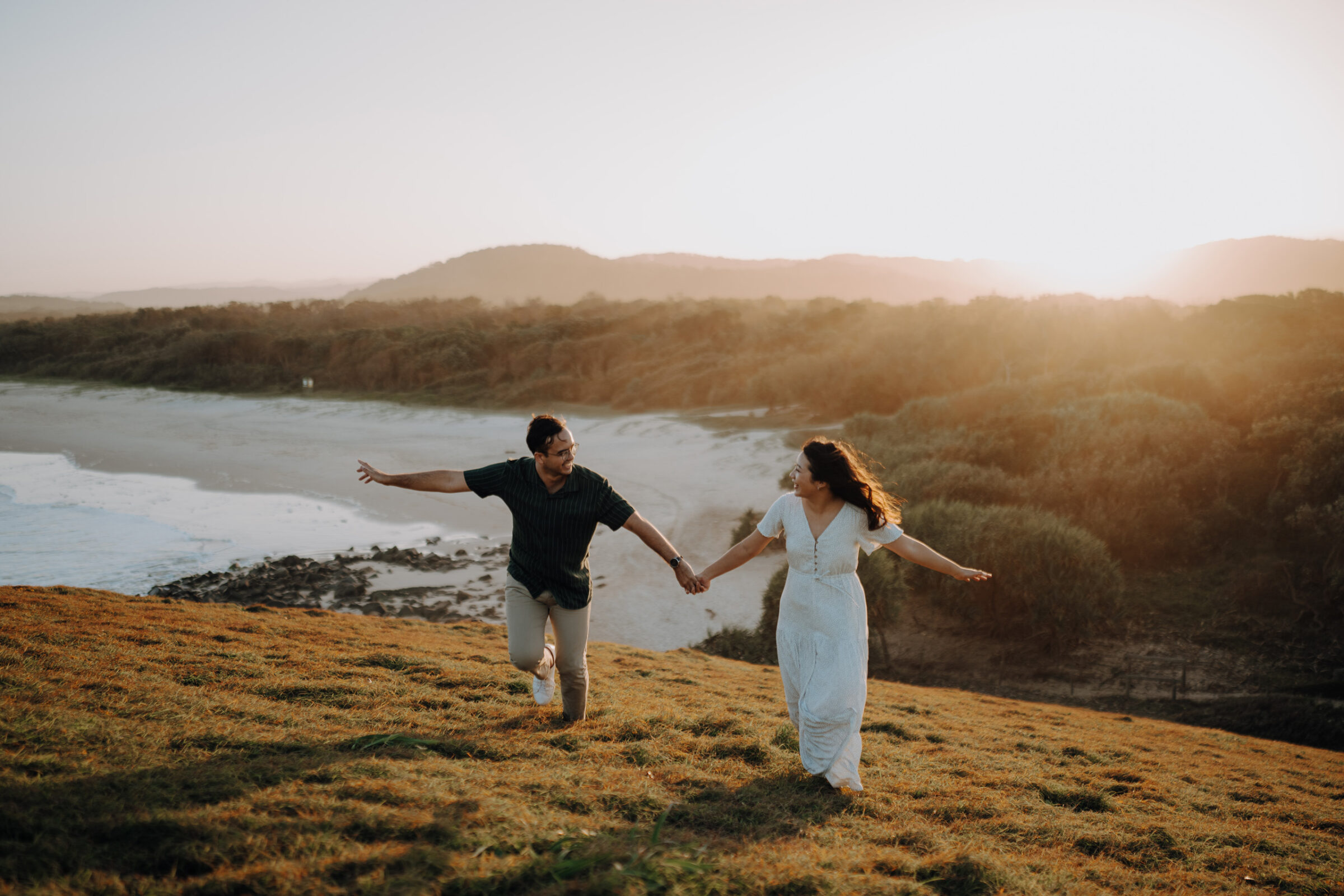 A couple joyfully runs hand-in-hand on a grassy hill by the coast, with the sun setting in the background.