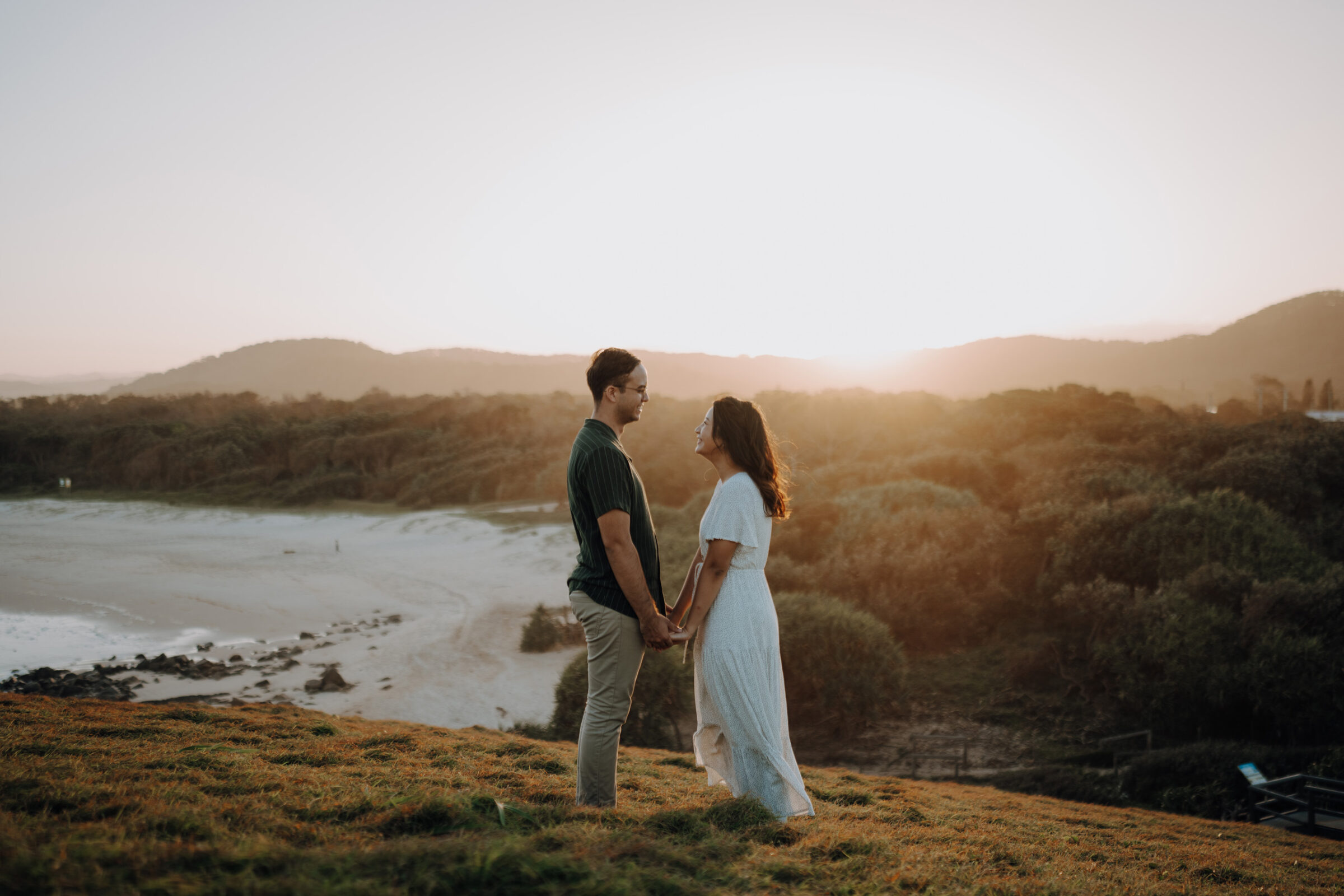 A couple stands holding hands on a grassy hill at sunset, with a beach and trees in the background.