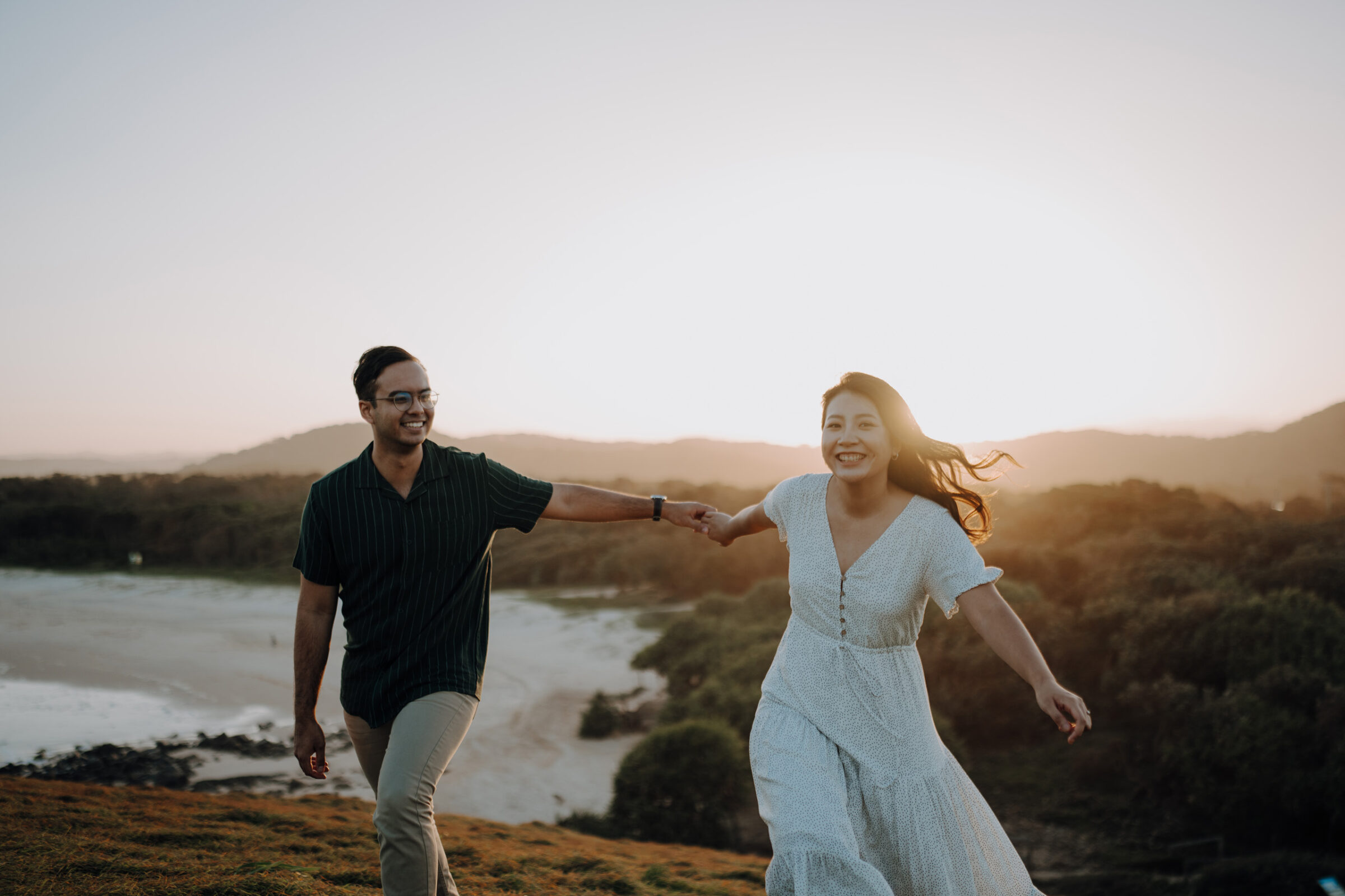 A couple holding hands and walking on a grassy hill at sunset with a beach in the background.