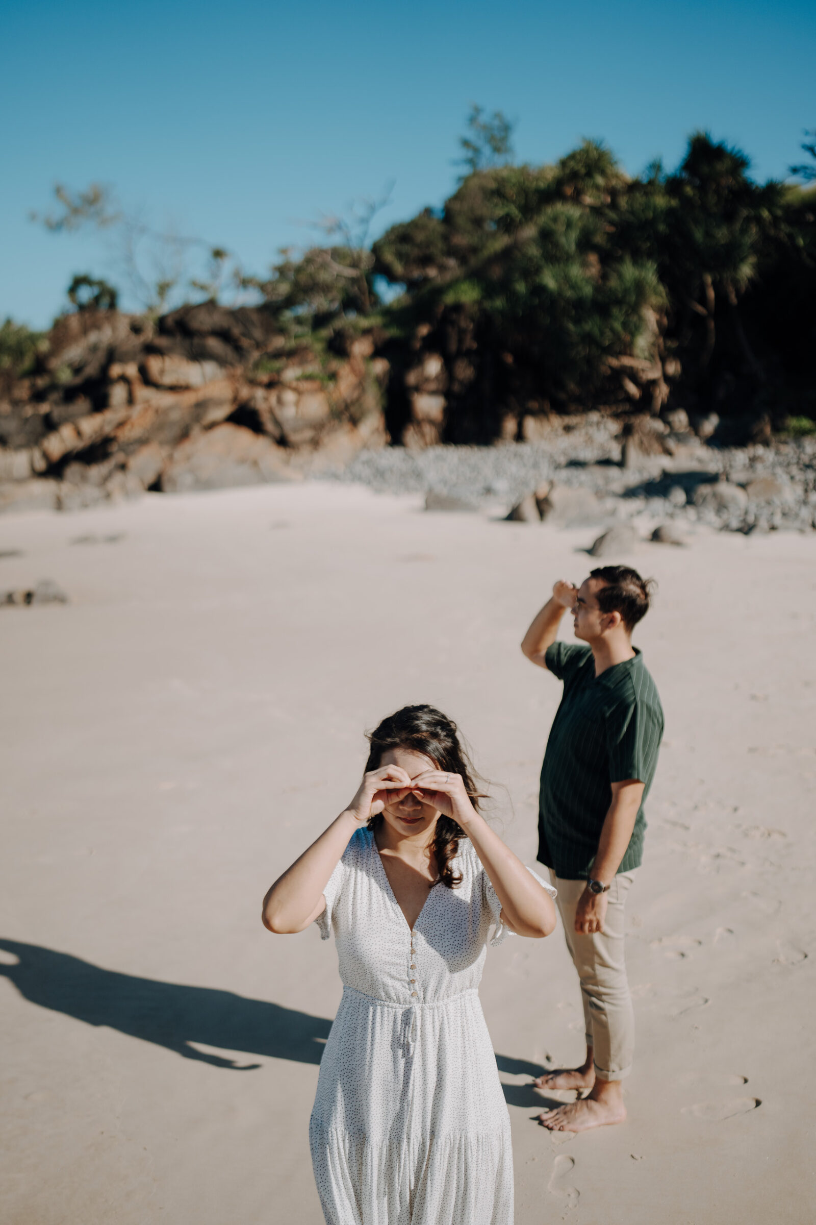 A woman in a white dress and a man in a green shirt stand on a sandy beach, using their hands to shade their eyes from the sun. Rocks and vegetation are visible in the background.