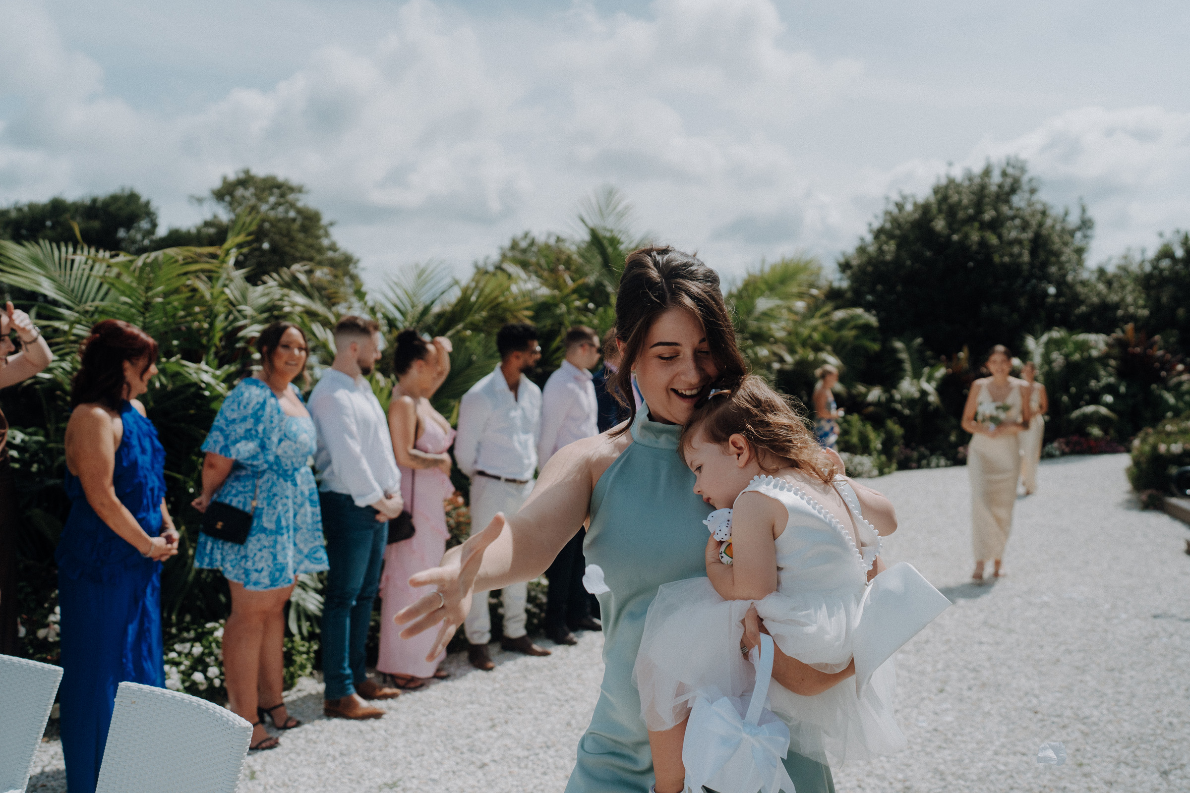 Woman in a blue dress holds a sleeping child in a white dress during an outdoor event. People in the background stand on a gravel path surrounded by greenery.