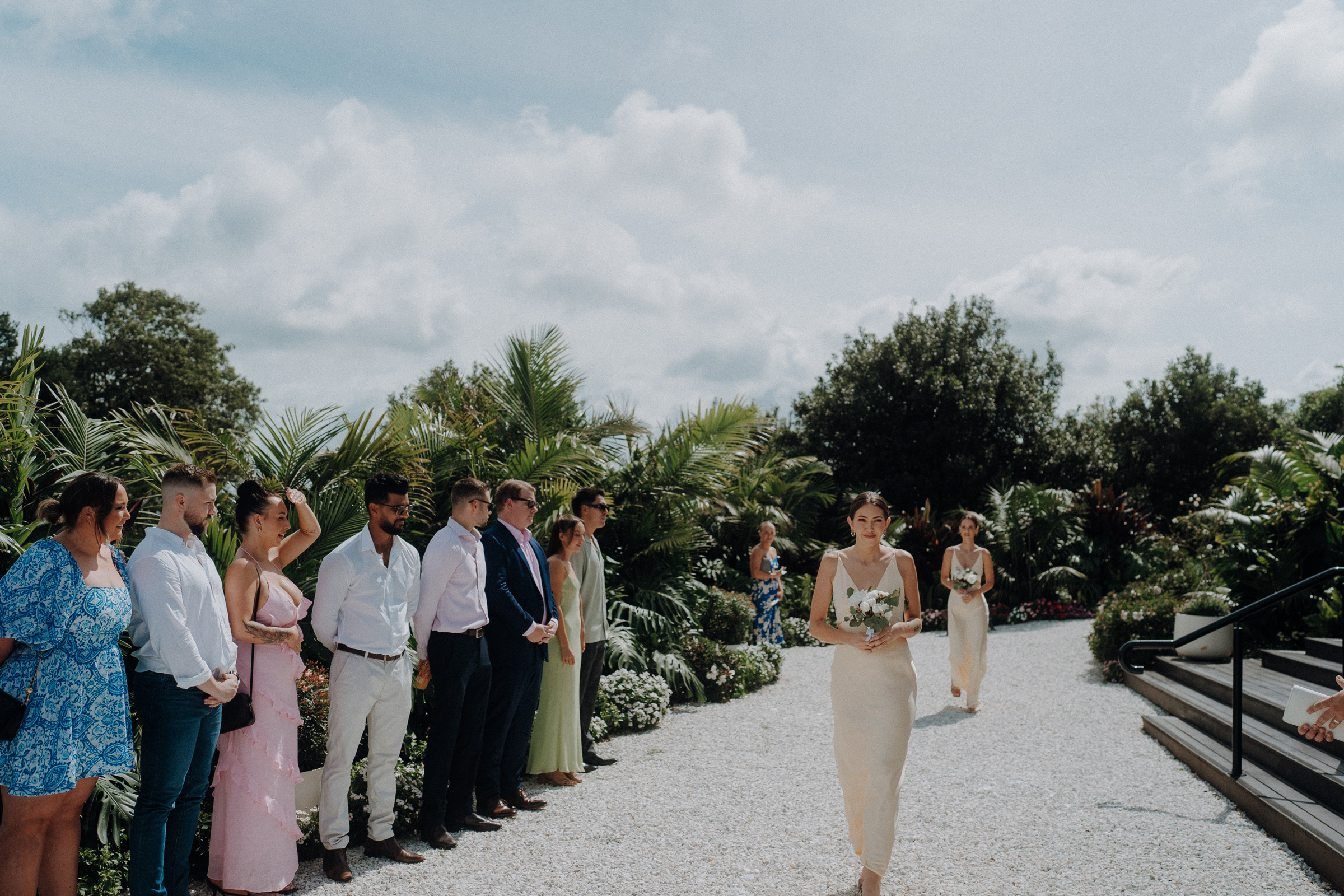 A bridesmaid walks down an outdoor aisle holding a bouquet, while guests stand on either side in a garden setting.