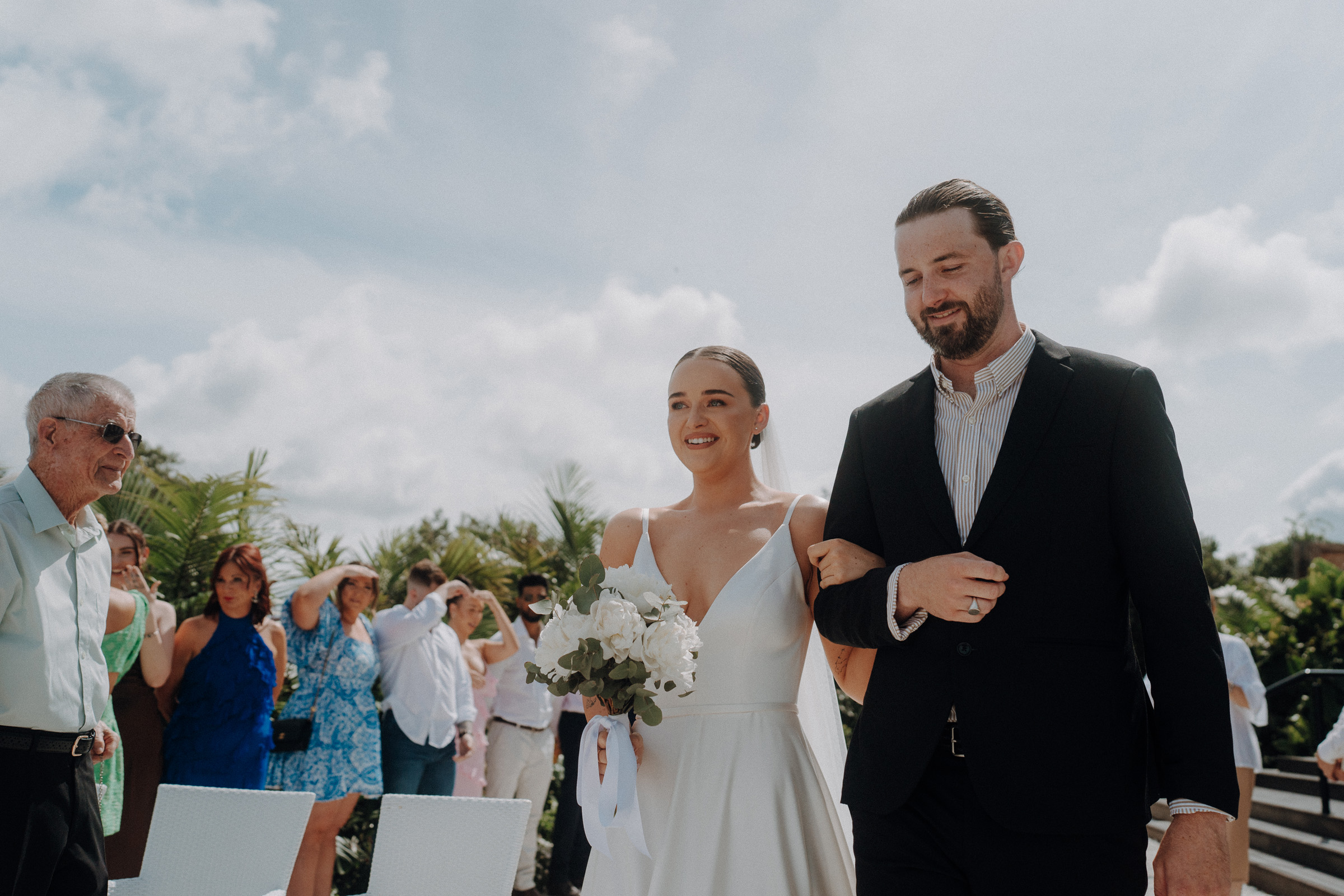 A bride holding a bouquet walks arm-in-arm with a man in a suit, while guests watch the outdoor ceremony.