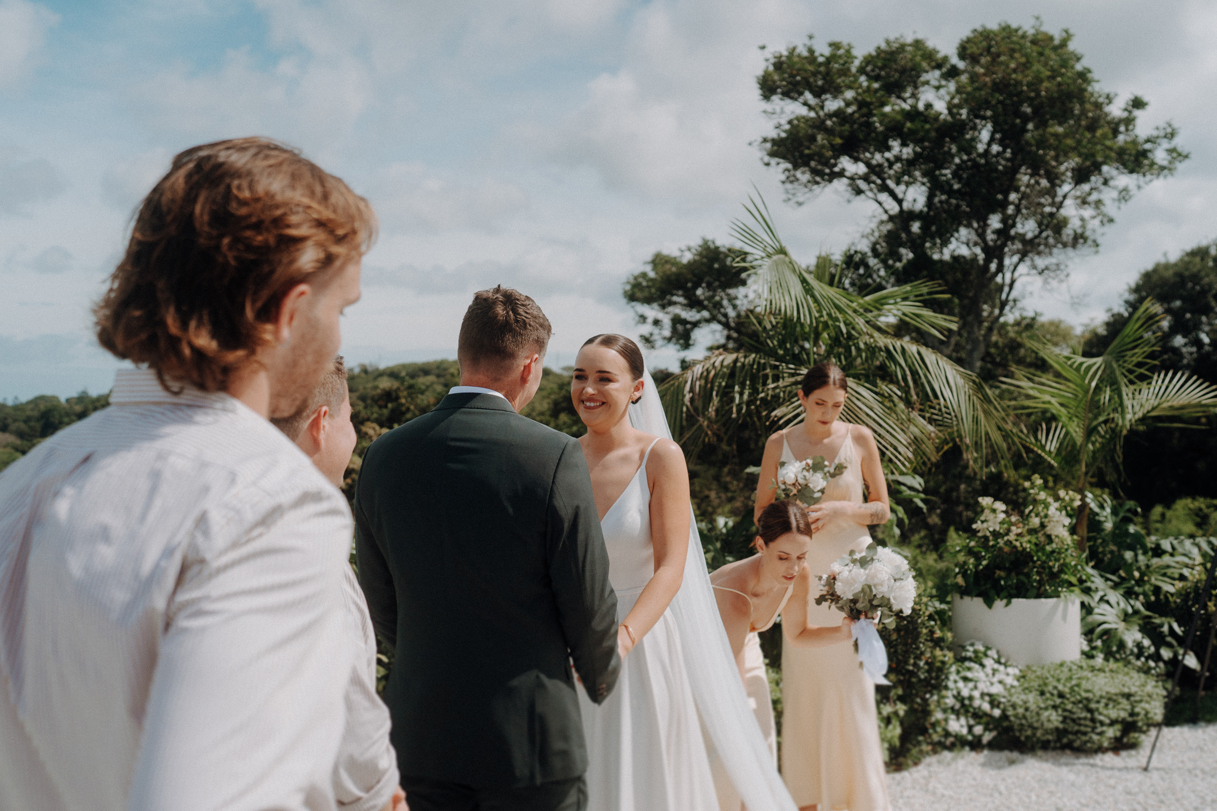 A bride and groom stand facing each other during a wedding ceremony outdoors, surrounded by bridesmaids holding bouquets.