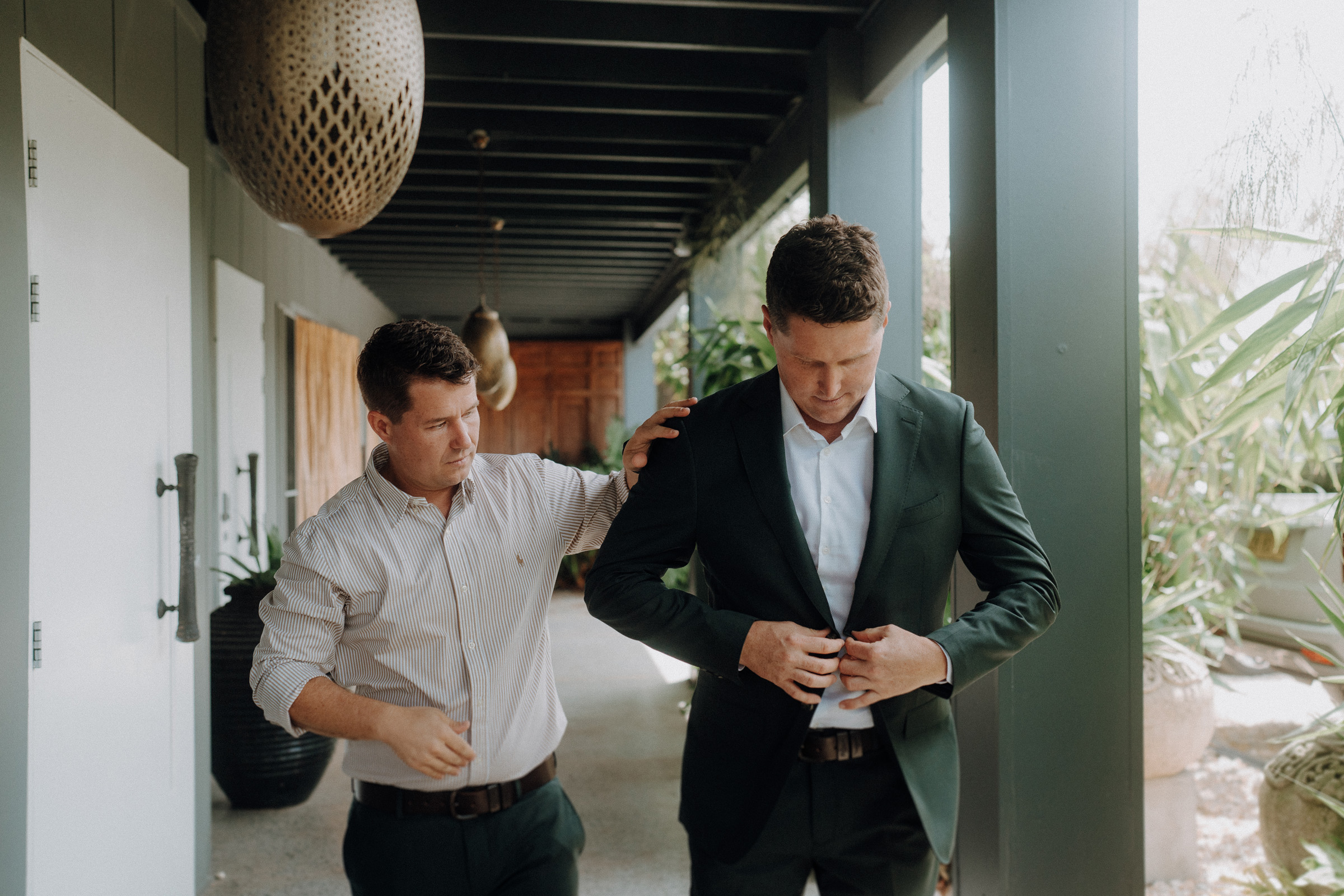 A man in a suit is buttoning his jacket while another man in a shirt assists him in a corridor with greenery and hanging decor.