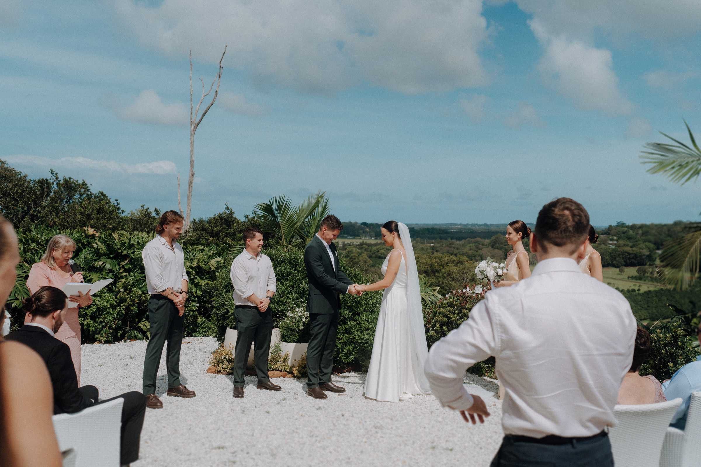 A couple stands holding hands during an outdoor wedding ceremony, surrounded by guests and lush greenery under a partly cloudy sky.