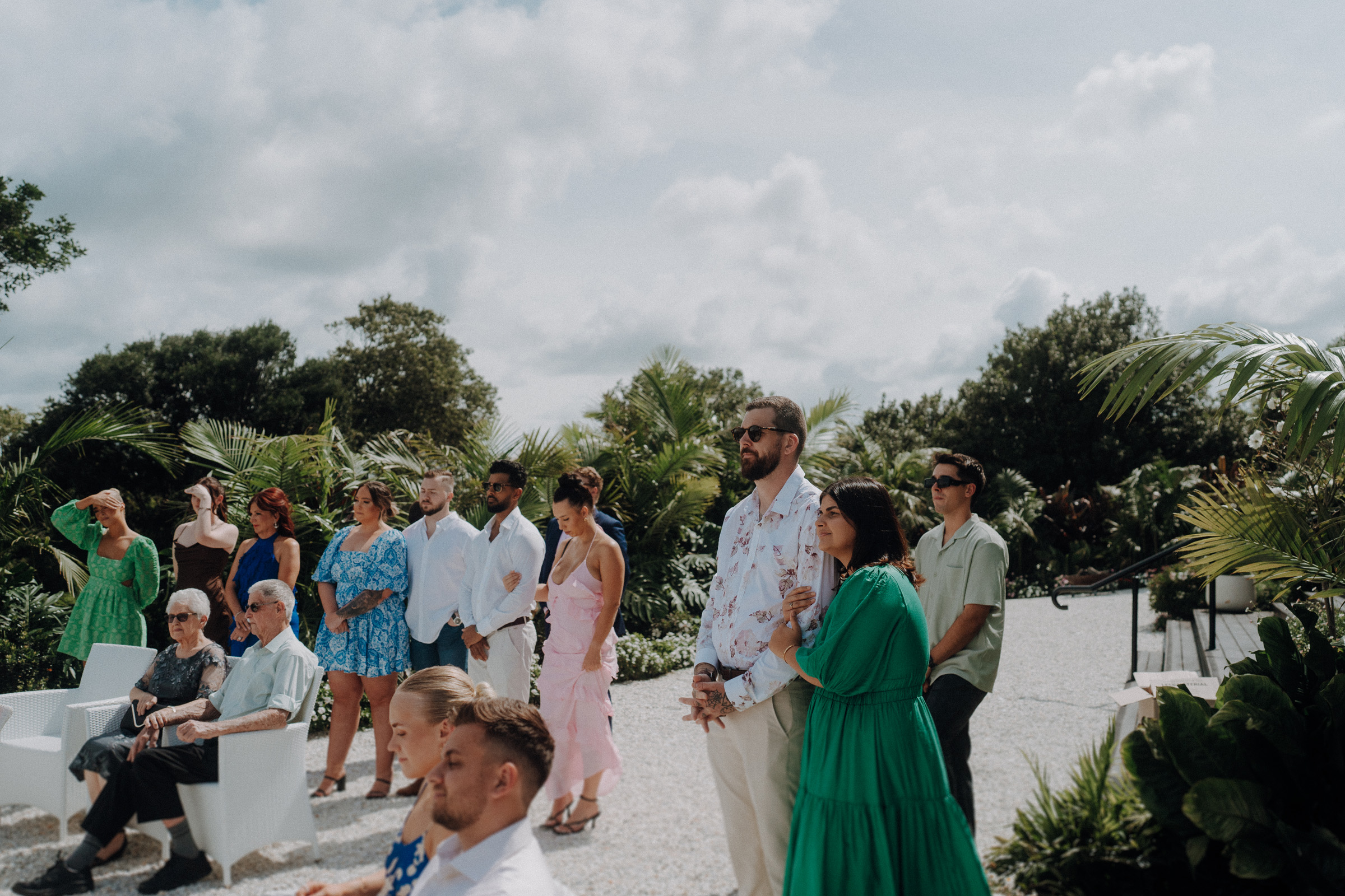 A group of people, including seated elderly individuals and standing adults in summer attire, gather outdoors on a sunny day amidst greenery.