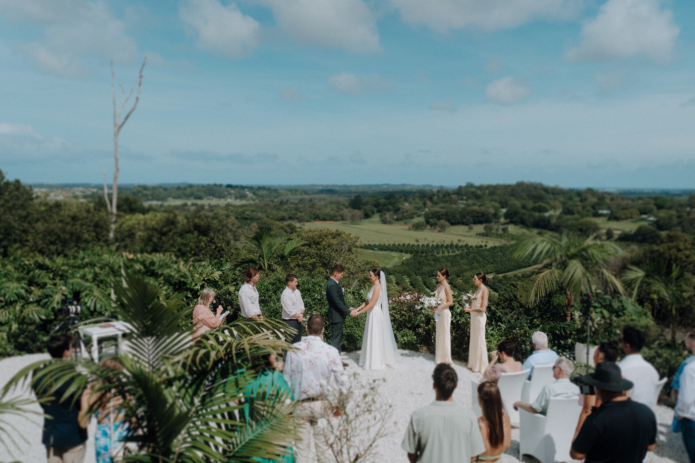 Outdoor wedding ceremony with a couple exchanging vows, surrounded by guests and lush greenery, under a clear blue sky.