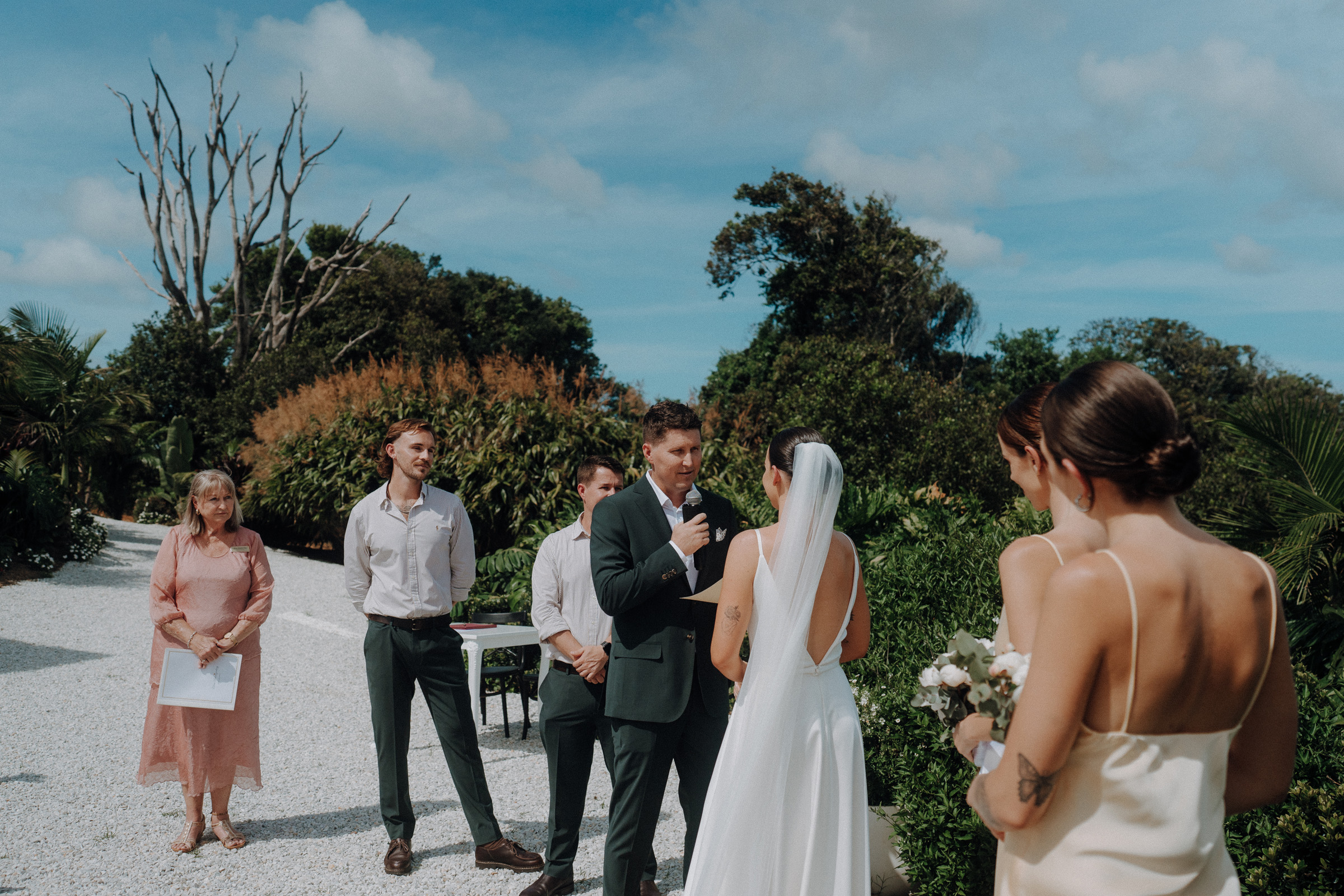 A couple exchanges vows outdoors, surrounded by wedding guests. The groom holds a microphone, and the bride wears a veil. Lush greenery and blue skies form the backdrop.