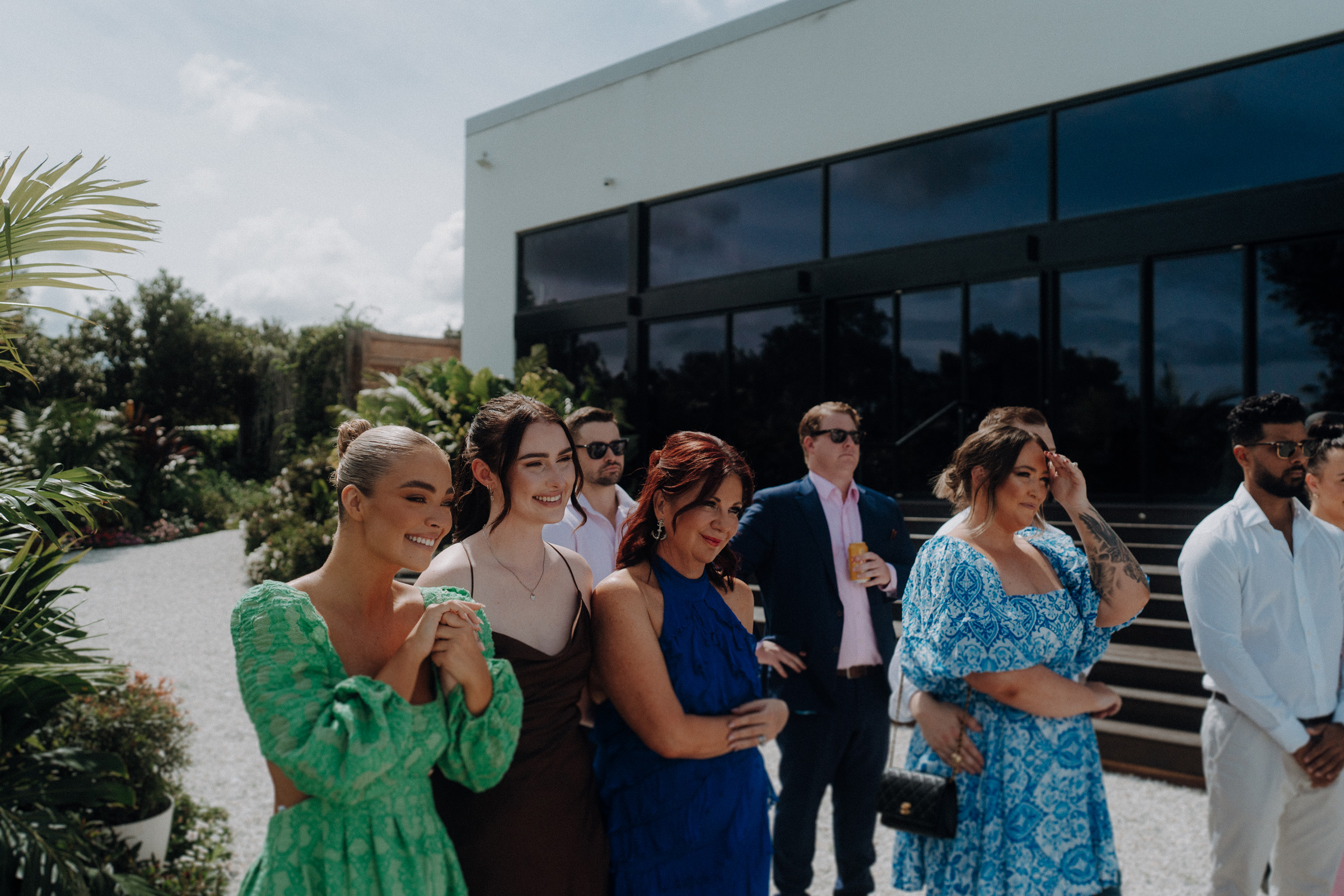 A group of people stands outside a modern building. They are dressed in formal attire, with some smiling and others watching intently. Lush greenery surrounds the area.