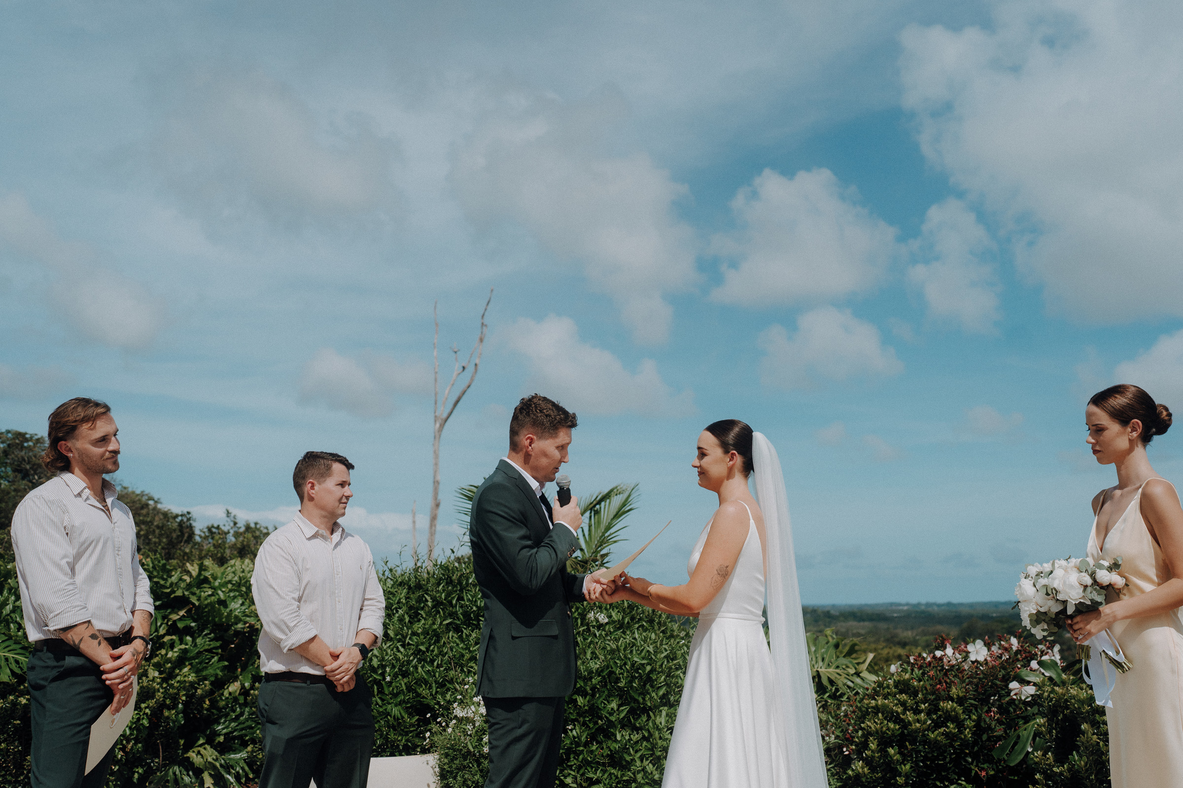 A couple exchanges vows outdoors, surrounded by three attendants. The sky is clear with scattered clouds. The bride holds a bouquet and wears a veil.