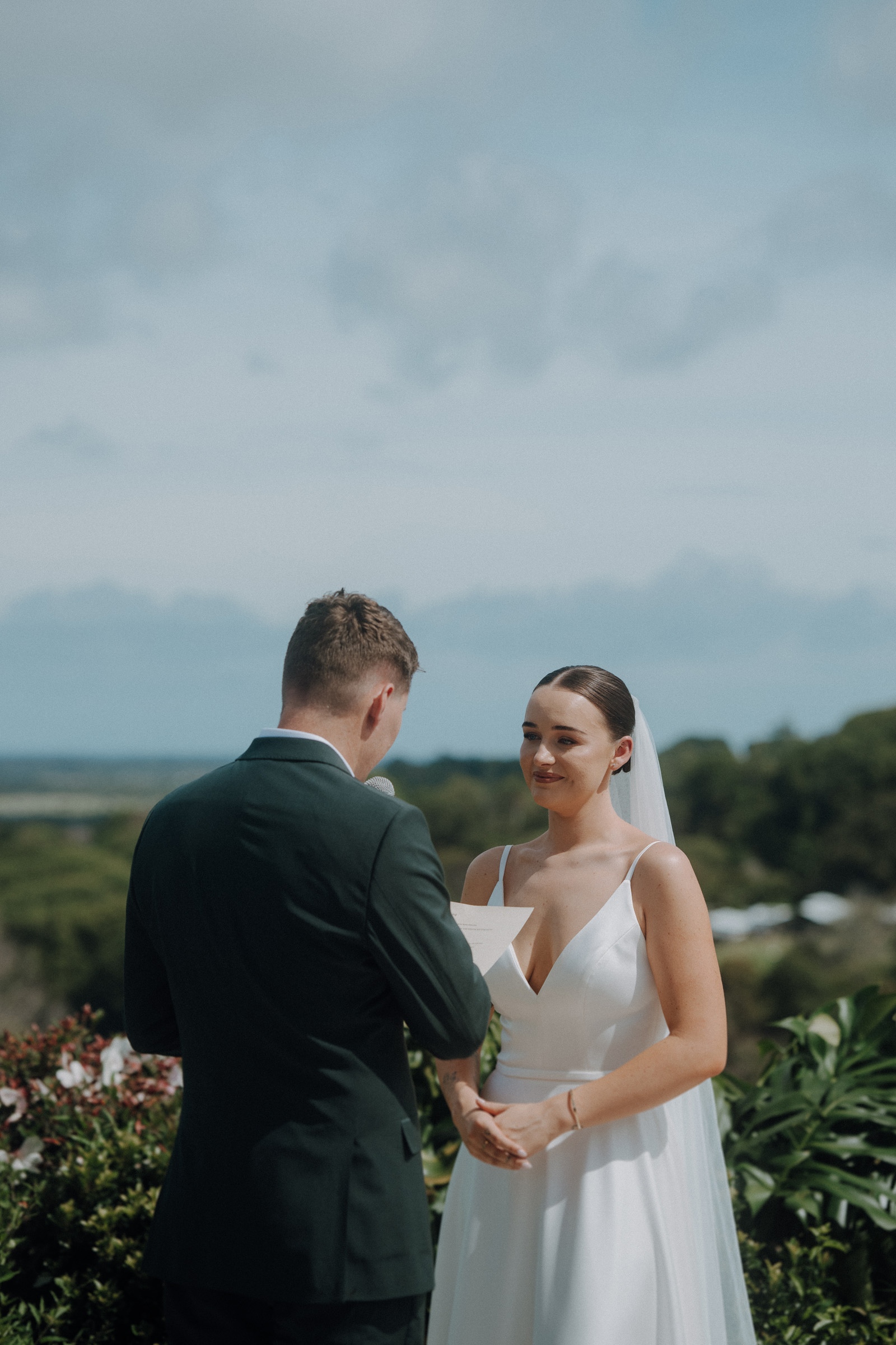 A couple holds hands outdoors during a wedding ceremony. The groom reads vows from a piece of paper while the bride listens intently.