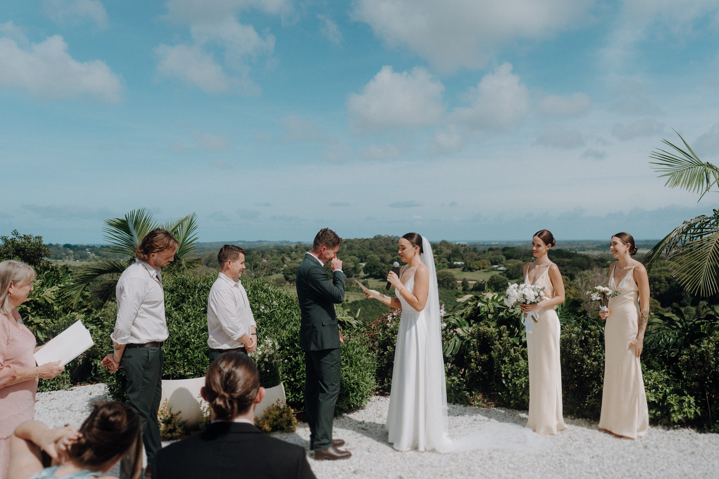 Outdoor wedding ceremony with a bride and groom exchanging vows. Bridesmaids in light dresses hold flowers. Guests and officiant stand nearby. Scenic landscape with greenery and blue sky in the background.