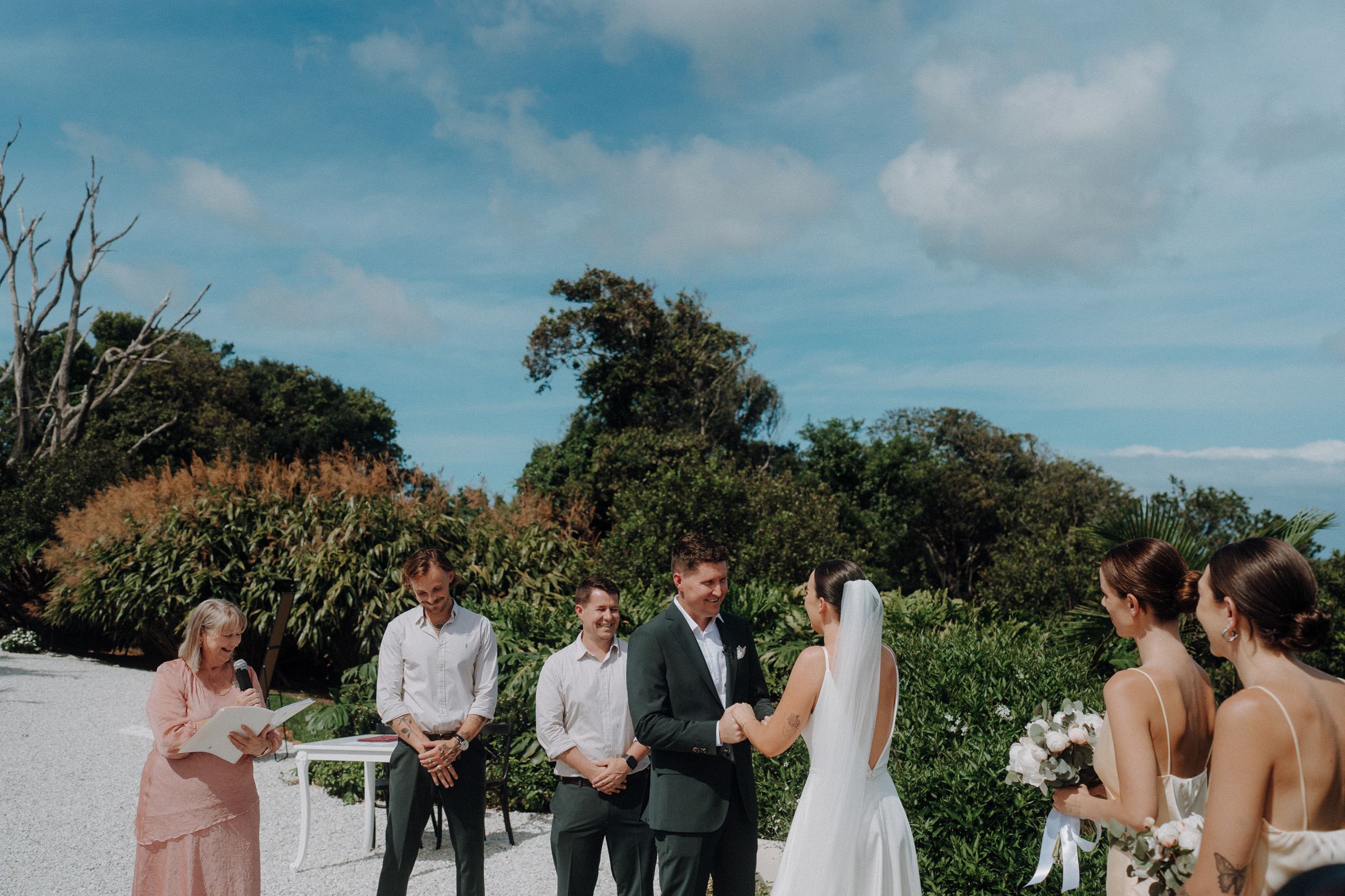 A couple exchanging vows outdoors, with an officiant and two men standing beside them. Two women holding flowers stand opposite. Trees and a bright sky serve as the backdrop.