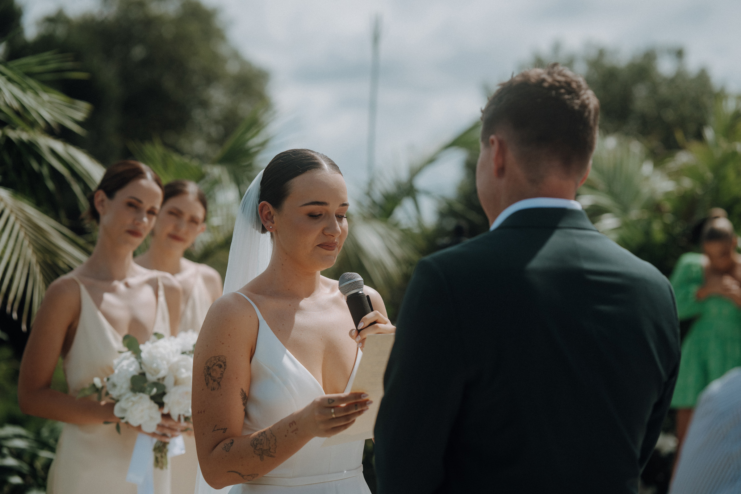 A bride reads vows from a paper to the groom outdoors, with bridesmaids holding flowers in the background.