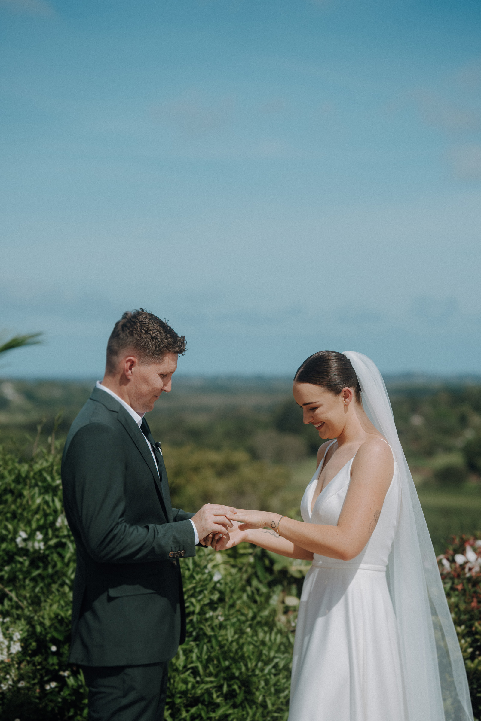 A bride and groom exchange rings outdoors on a sunny day, surrounded by greenery.