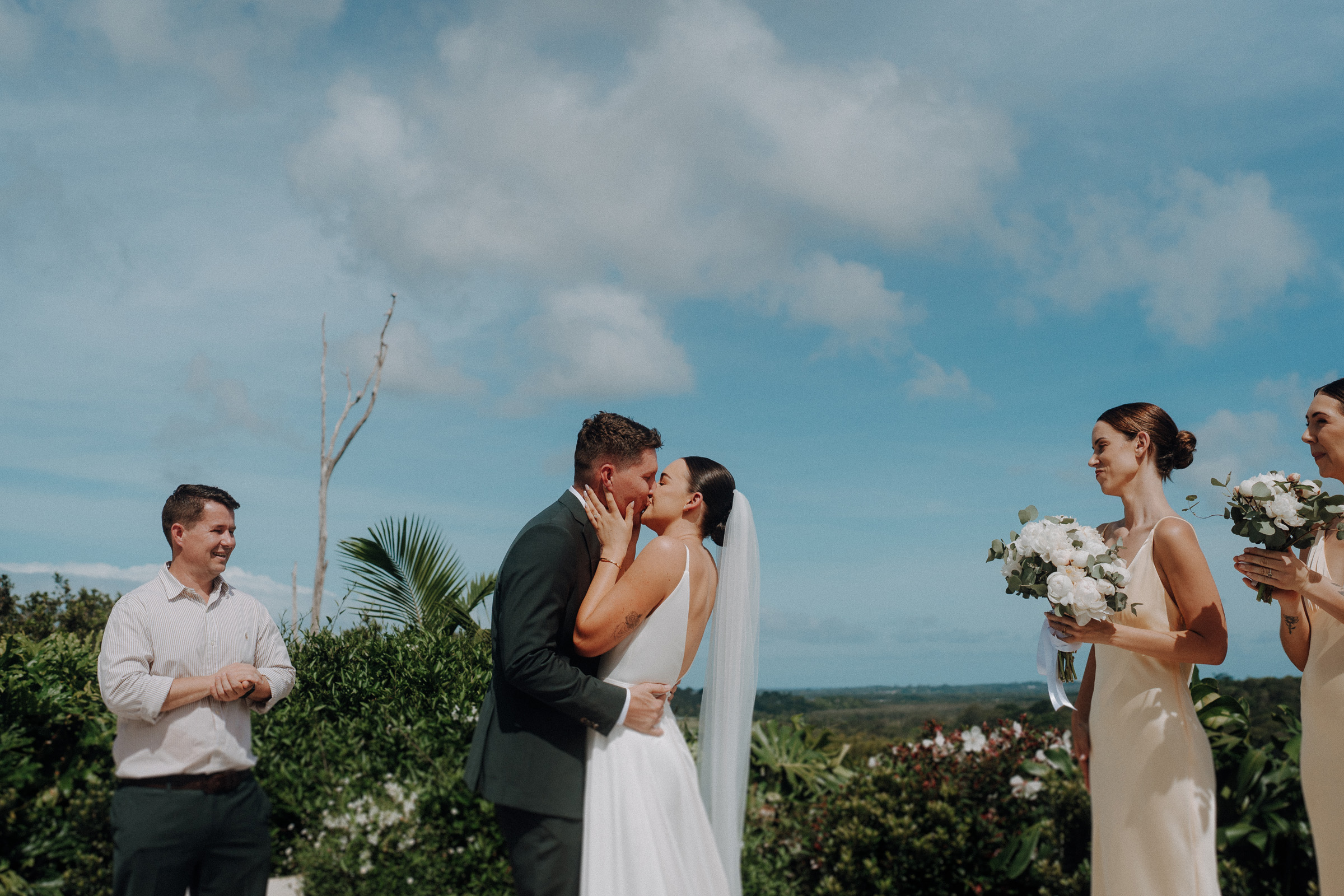 A bride and groom kiss at an outdoor wedding ceremony, with bridesmaids holding bouquets, set against a scenic backdrop of greenery and blue sky.