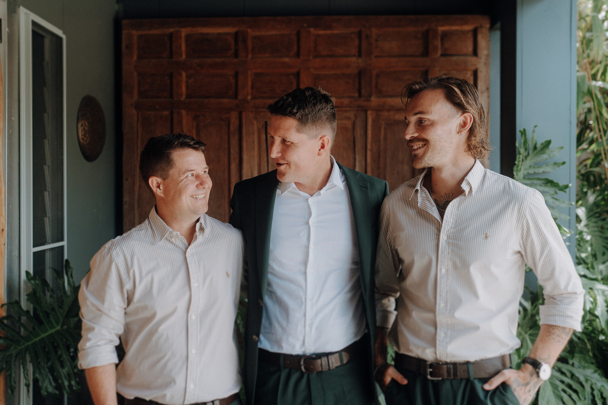 Three men in dress shirts and blazers stand smiling, with one in the center placing his arms around the others. They are in front of a wooden door with plants around them.