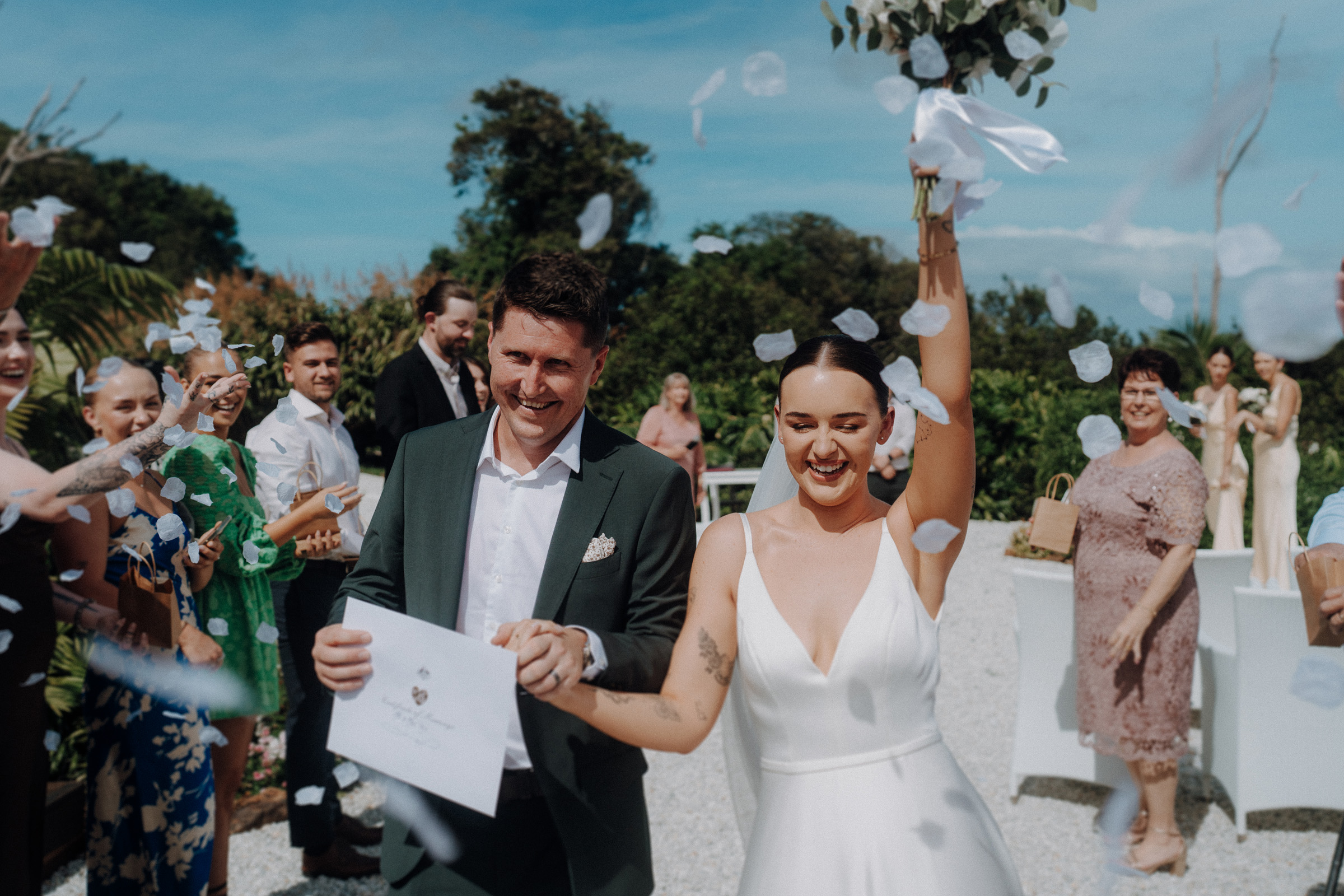 A bride and groom celebrate outside, surrounded by cheering guests. The bride holds a bouquet and raises her arm, while the groom holds a certificate. Petals are falling in the air.