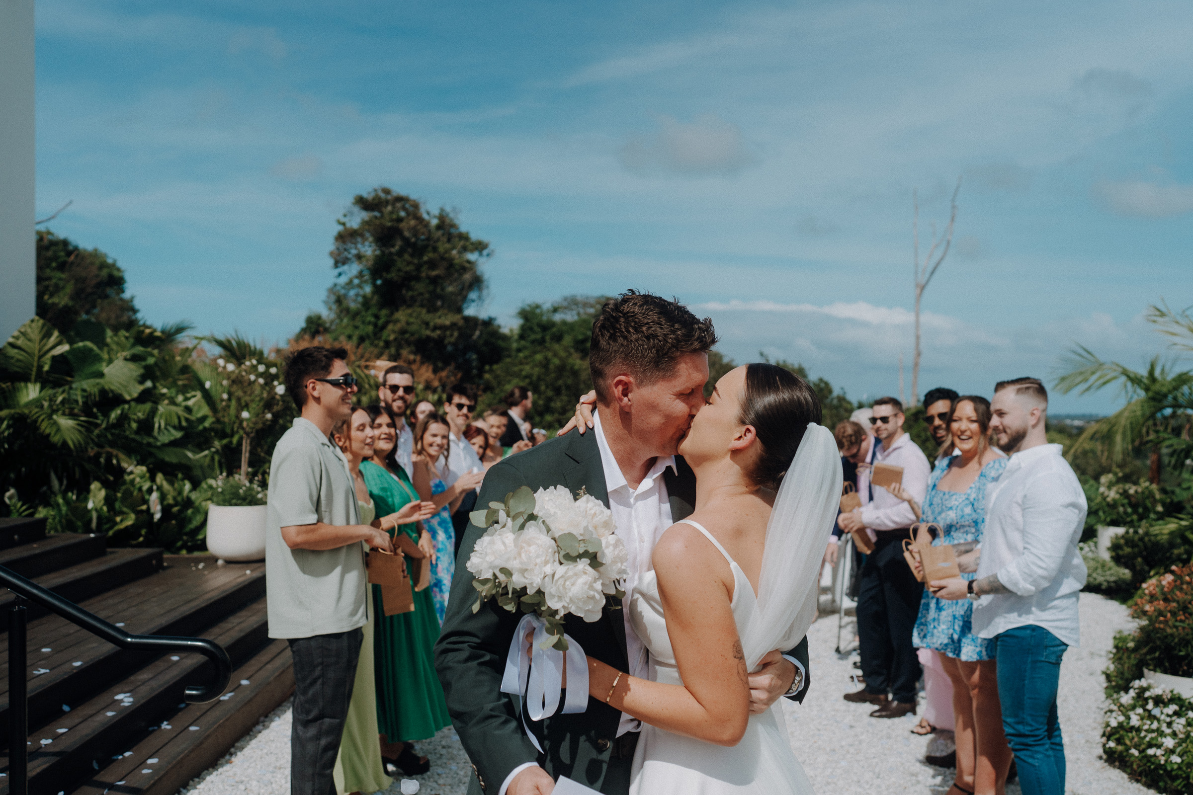 A bride and groom kiss at an outdoor wedding ceremony, surrounded by guests. The bride holds a bouquet of white flowers.