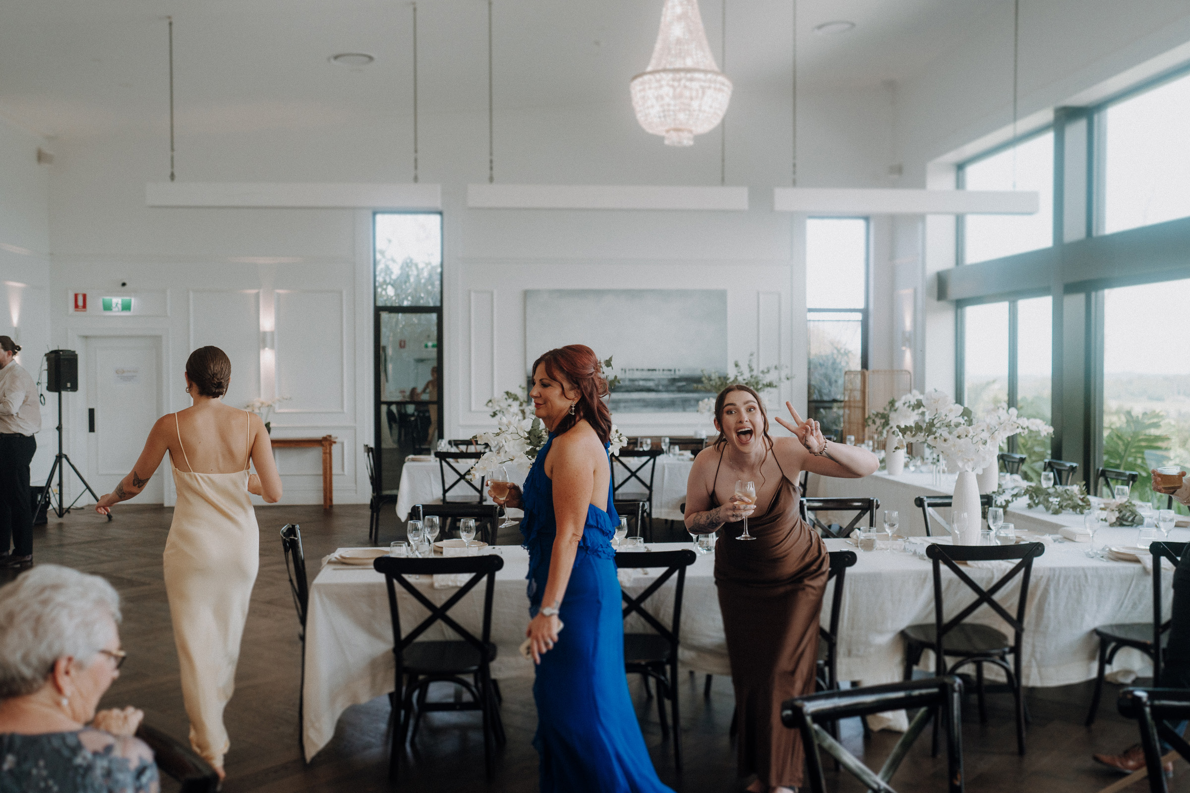 Three people in a decorated dining room; one poses playfully, another walks away. Tables are set with white flowers under a chandelier.