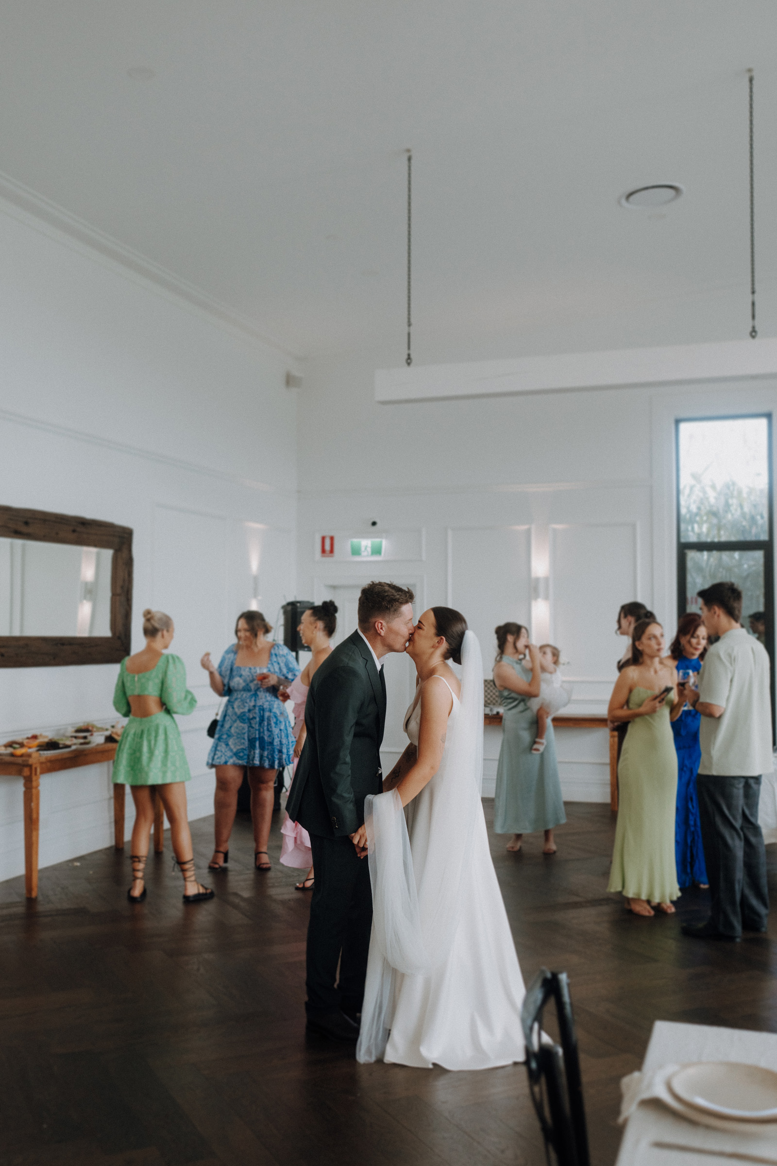 A bride and groom share a kiss in a reception hall. Guests in colorful dresses stand in the background, talking and holding beverages.