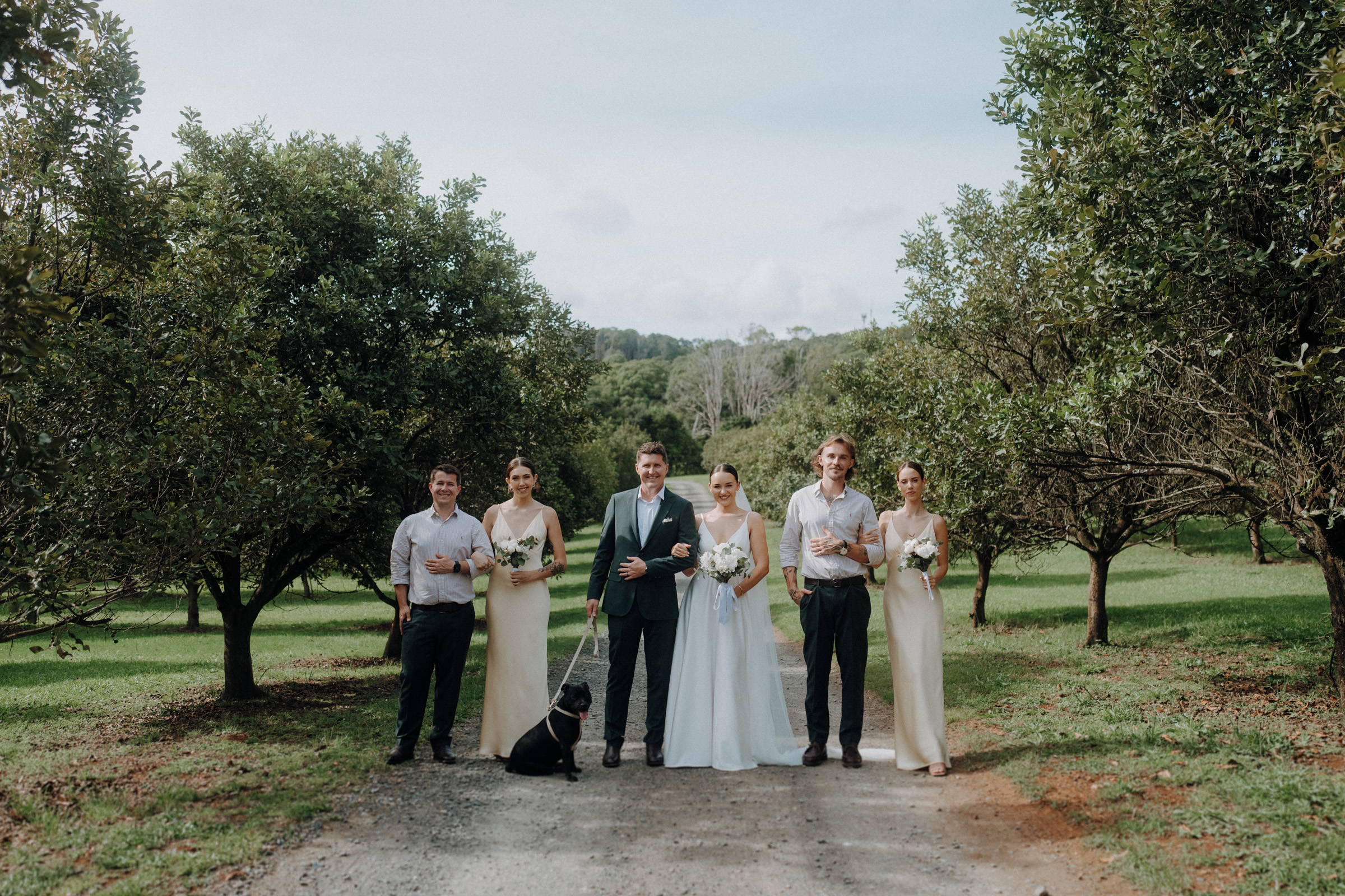 A bride and groom stand in the center of a small wedding group outdoors, with bridesmaids and groomsmen surrounding them. A black dog sits in front. Trees and greenery are in the background.