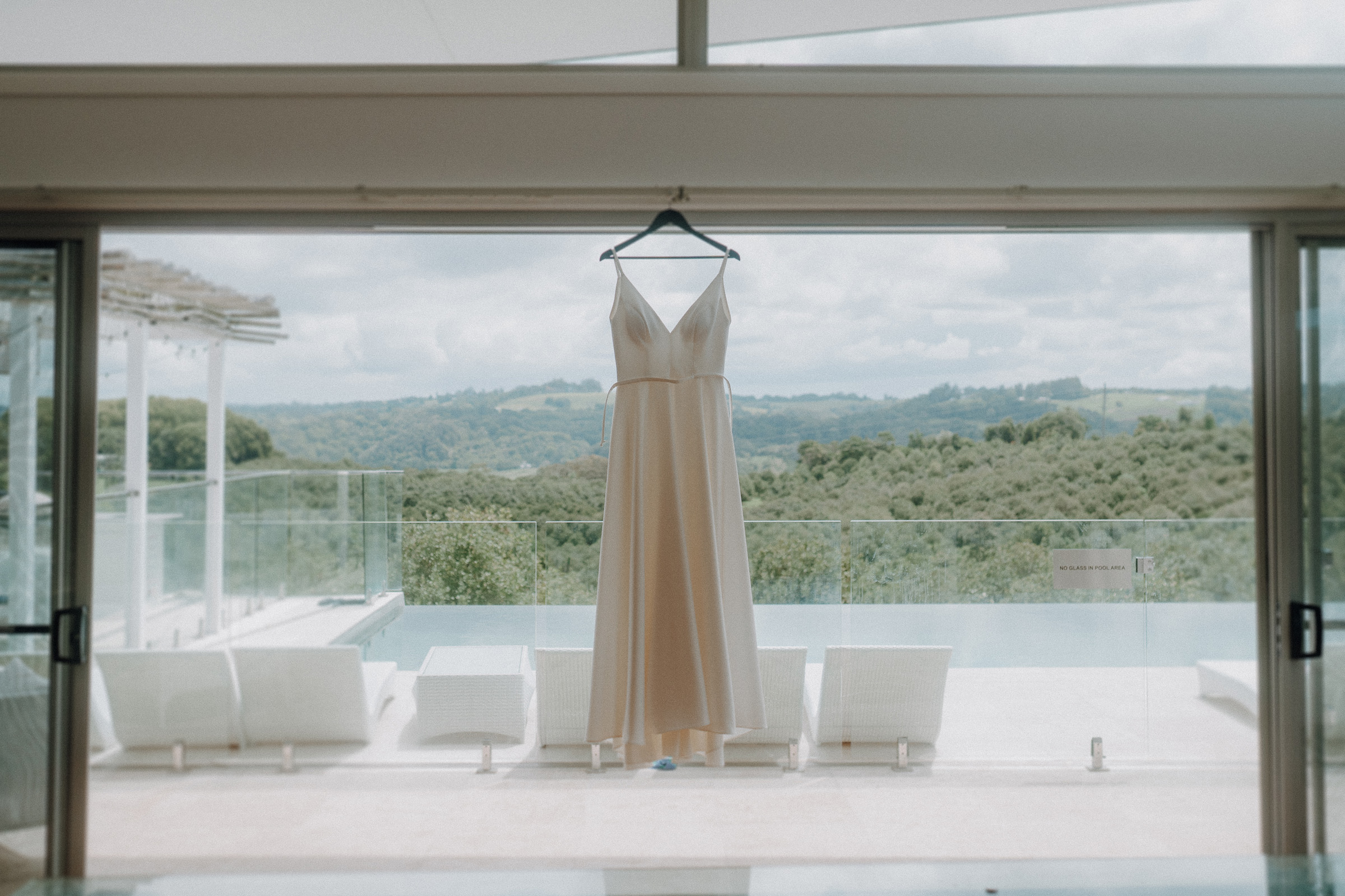 A white dress hangs in front of a large window overlooking a green landscape and a swimming pool.