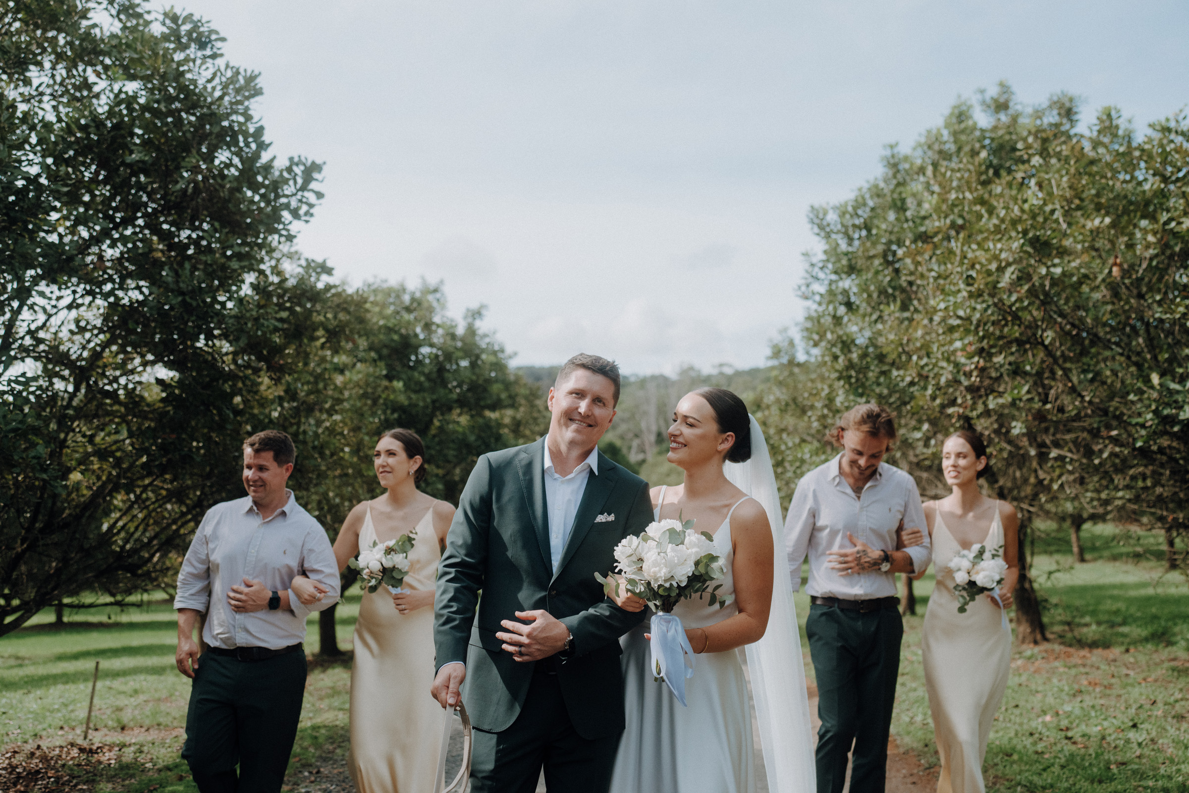 A bride and groom walk together in a garden setting, accompanied by four smiling attendants holding bouquets.