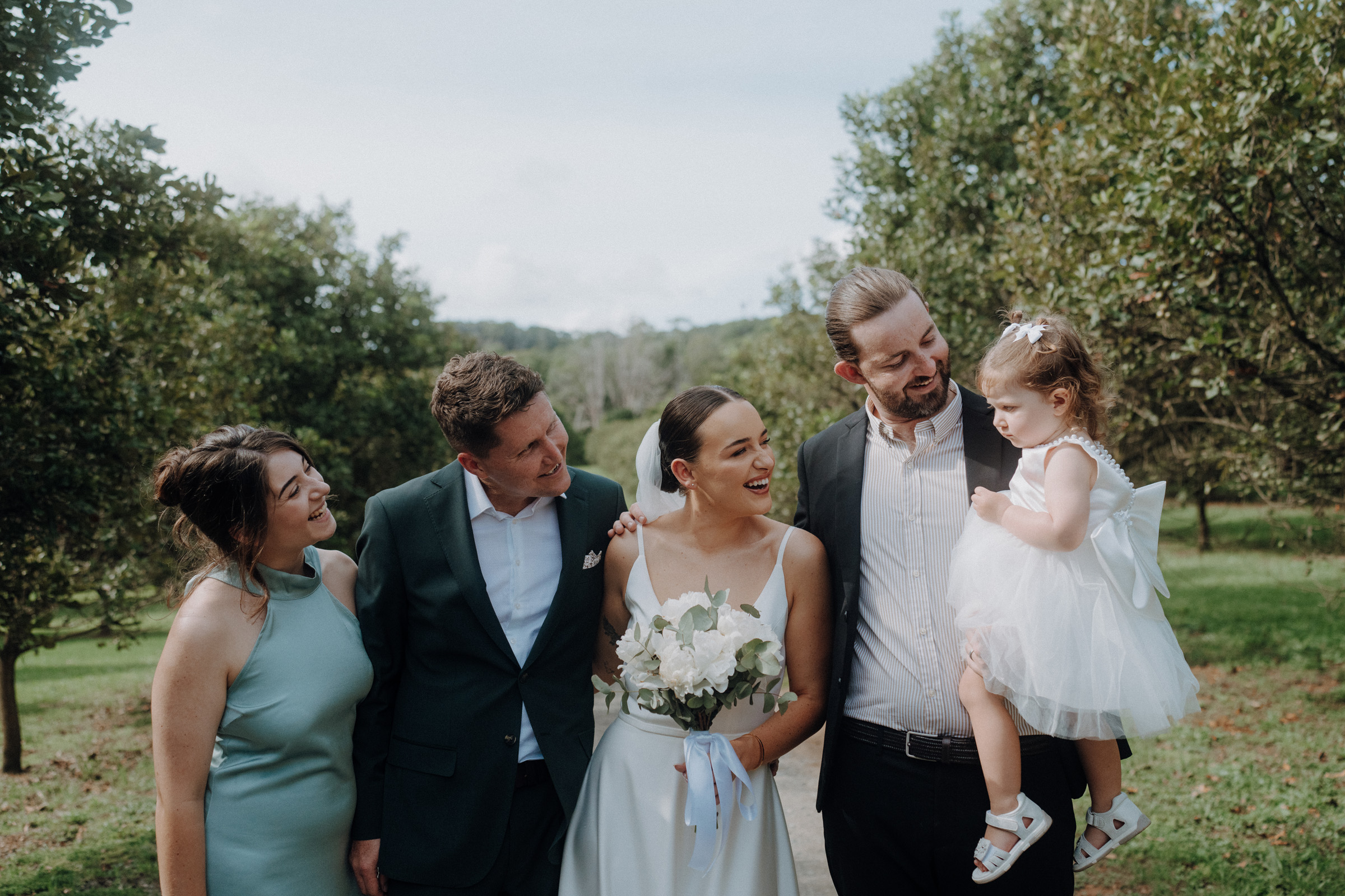 A newlywed couple stands outdoors with two other adults and a child in a white dress. They are smiling and looking at each other amidst greenery.