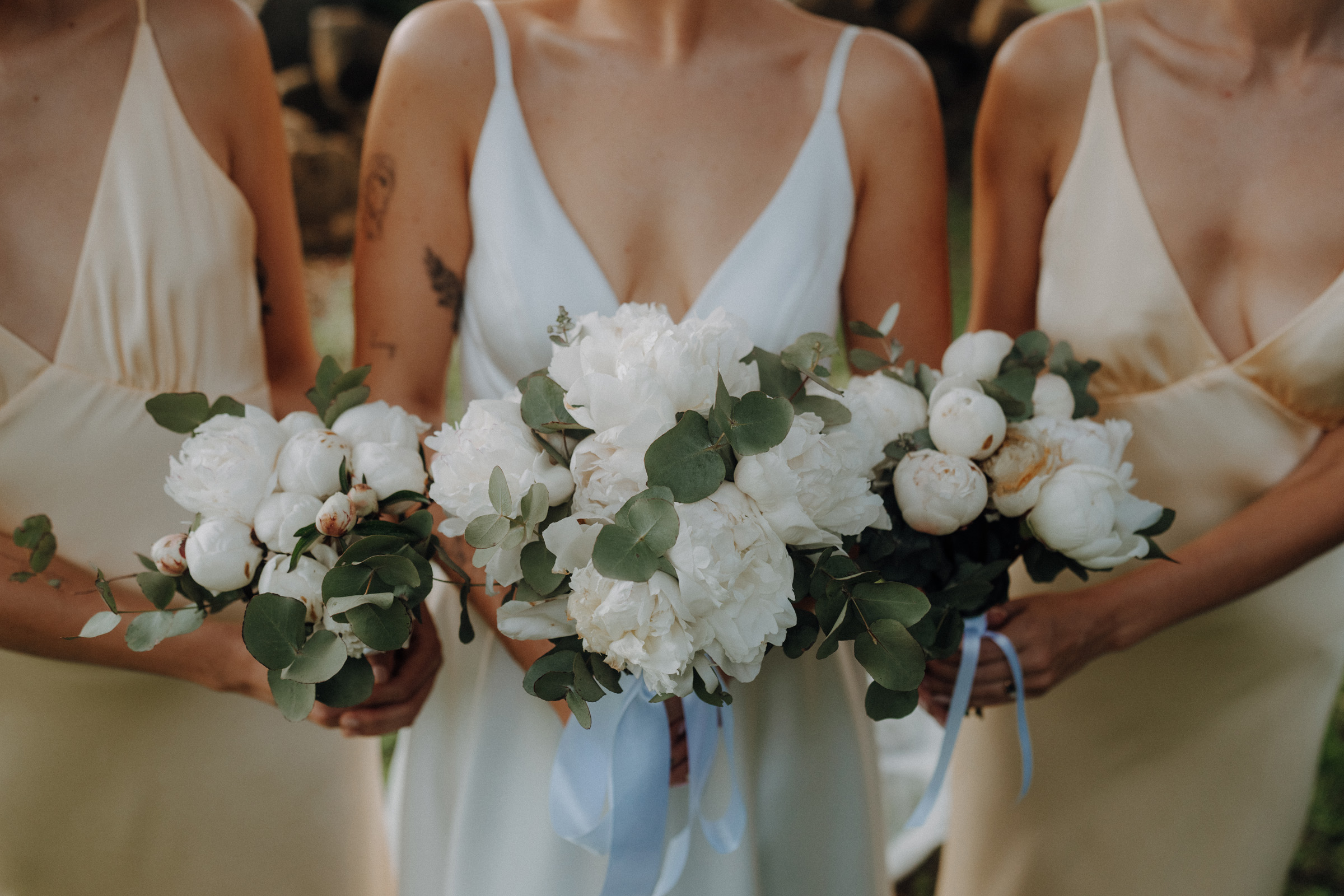 Three women holding white peony bouquets, dressed in light-colored gowns with V-necklines.