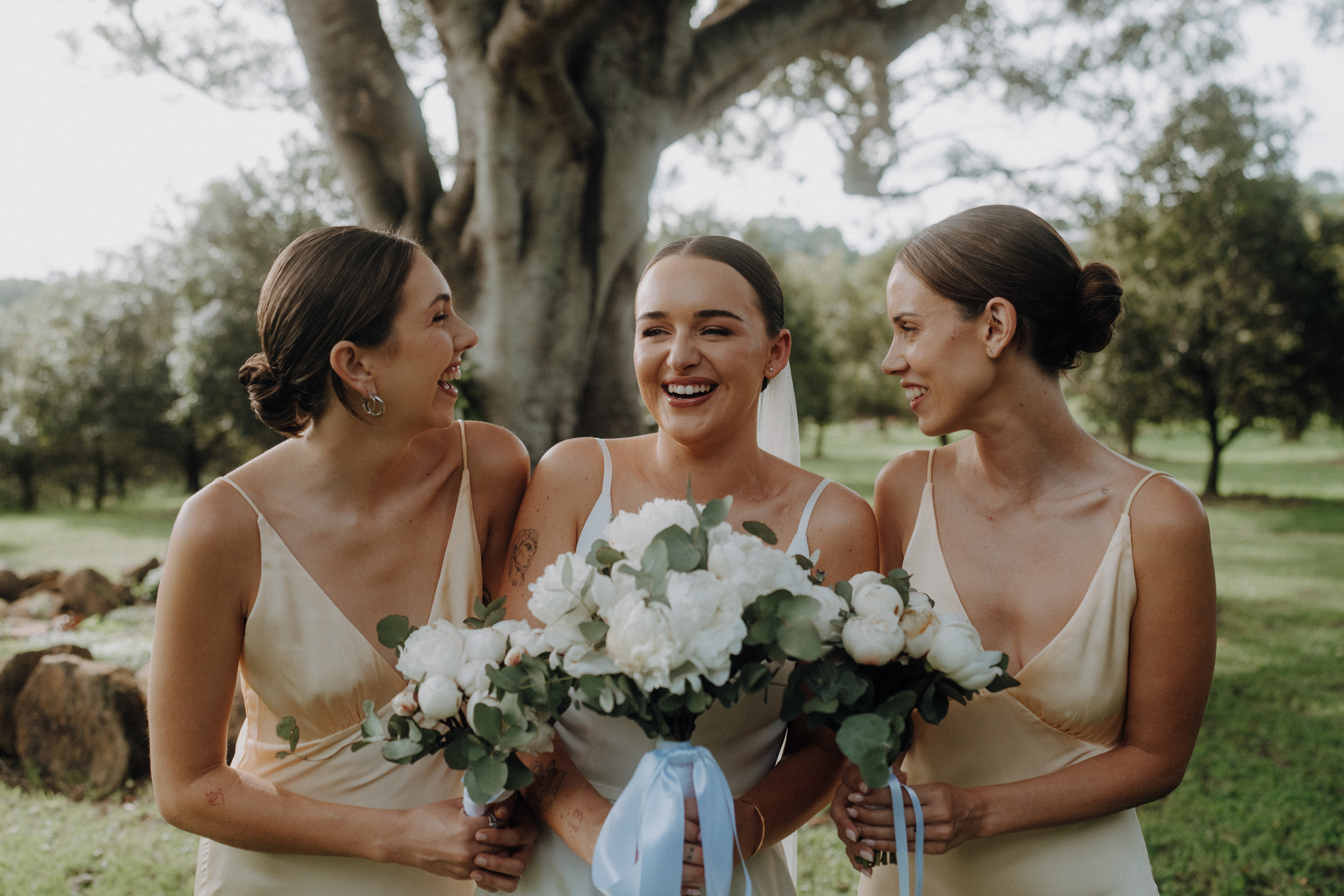 Three women in matching dresses laugh and hold flower bouquets in front of a large tree outdoors.