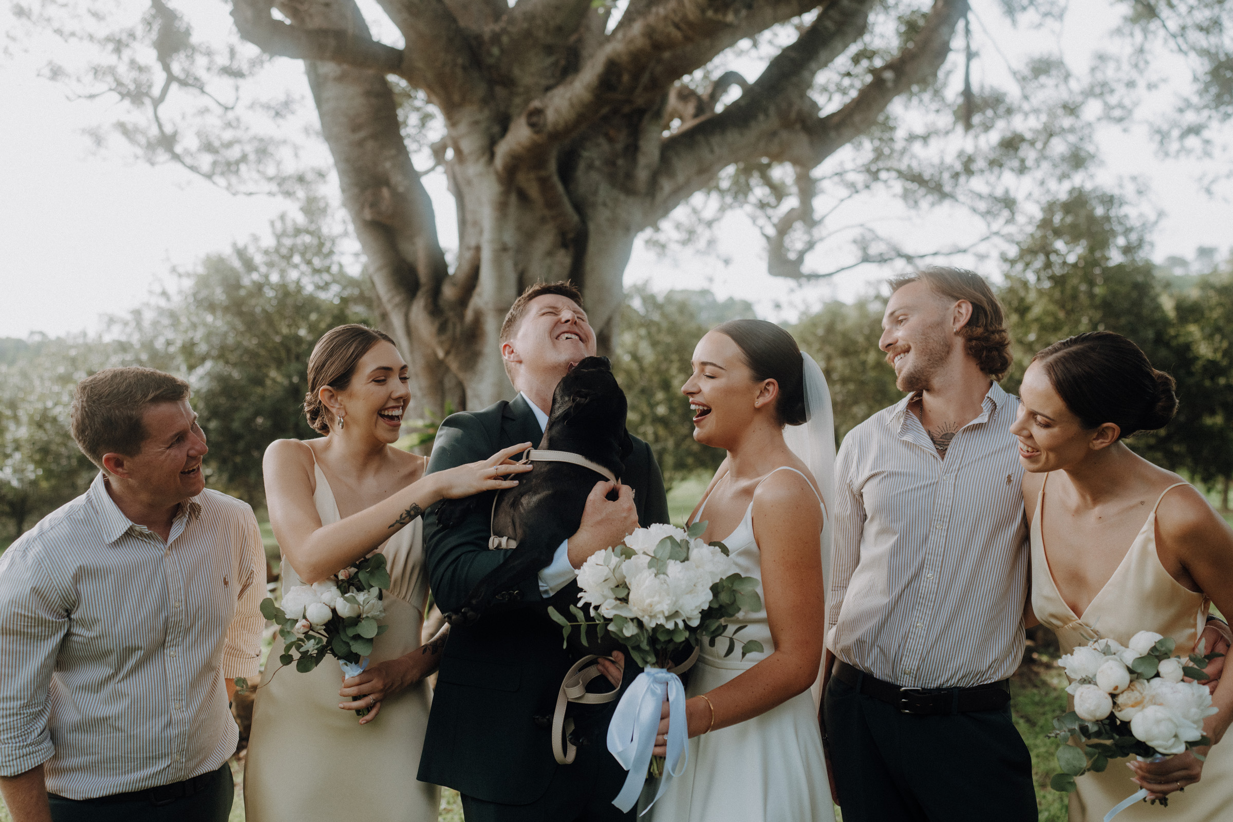 Wedding party laughing with a couple holding a small dog; three women in light dresses and two men in shirts stand in a garden setting.