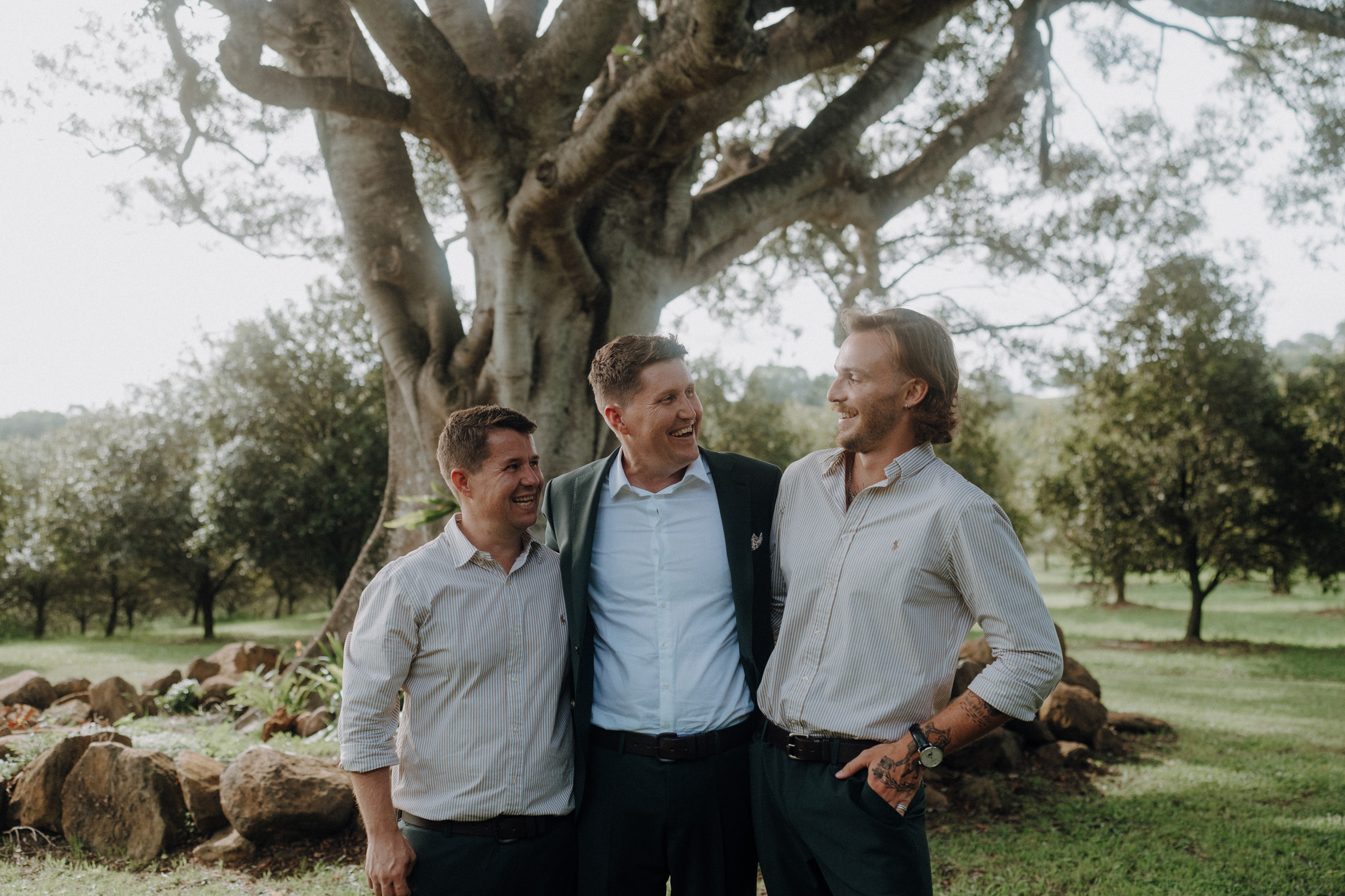 Three men stand together smiling under a large tree in a grassy area, with rocks and foliage in the background.