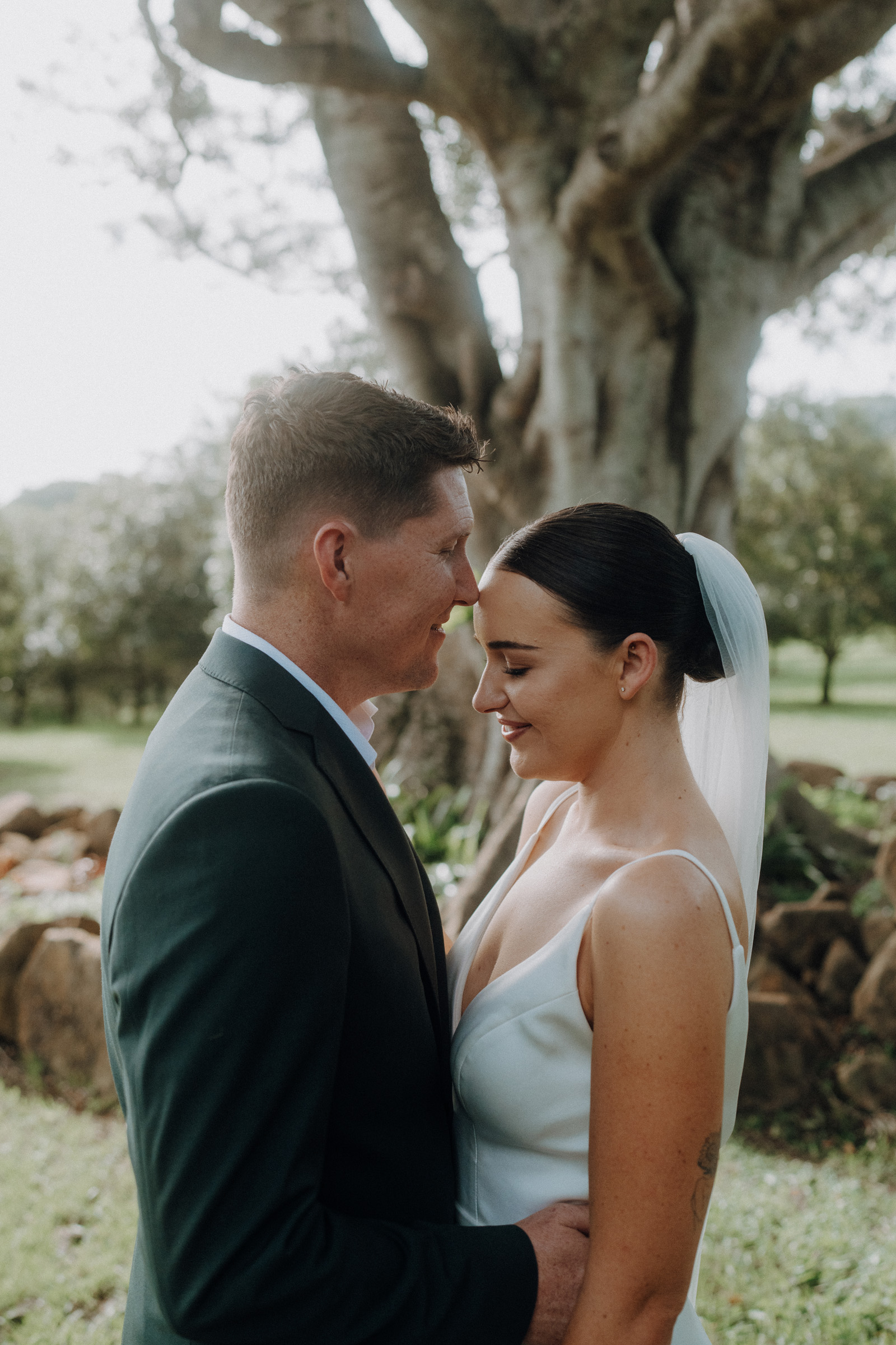 A couple stands close together, outdoors, beside a large tree, both dressed in formal attire with the woman in a white gown and veil.