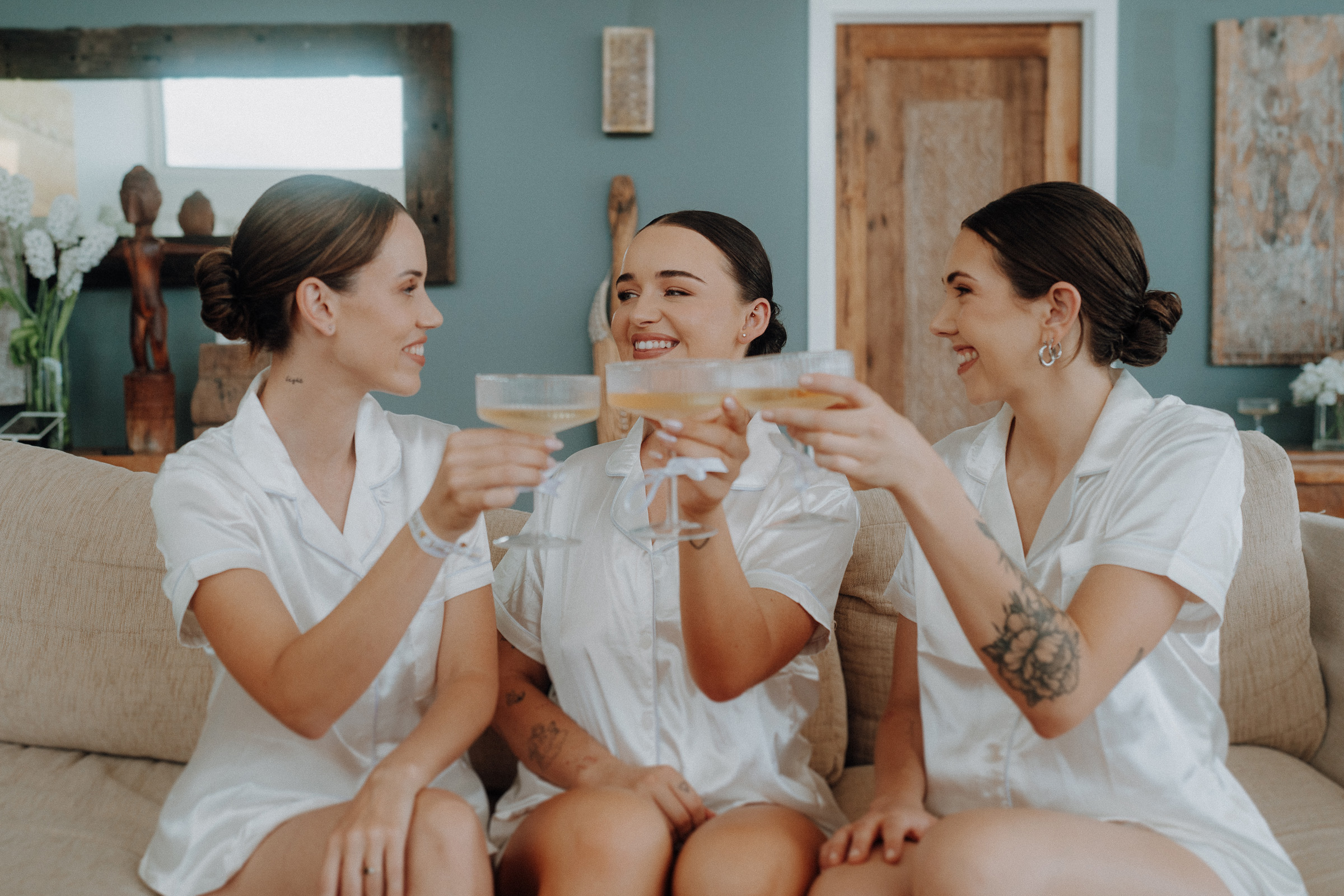 Three women in matching white outfits sit on a couch, smiling and holding champagne glasses, in a room with blue walls and wooden decor.