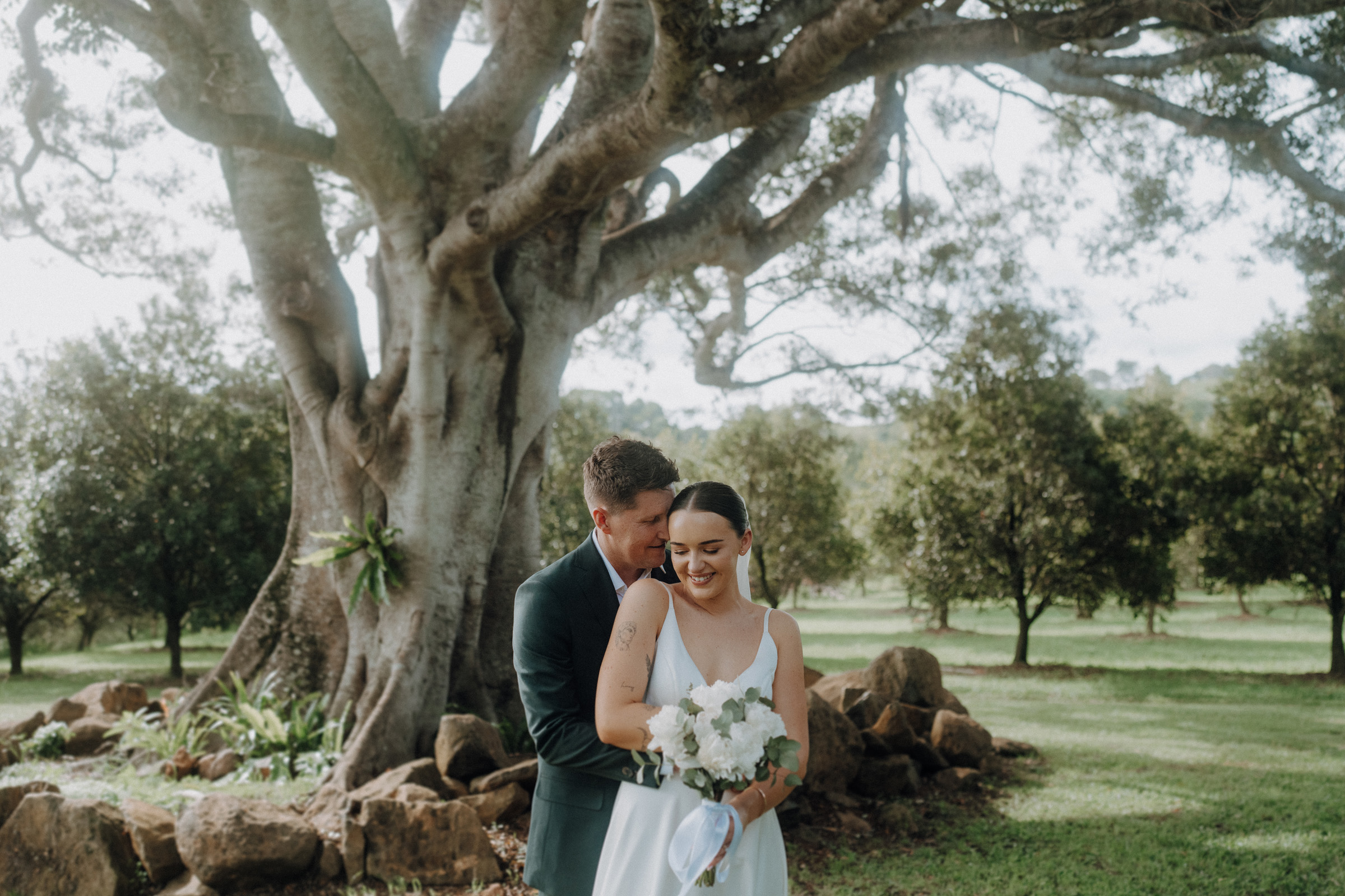 A couple in wedding attire stands closely together under a large tree in a garden, with the groom embracing the bride, who holds a bouquet of white flowers.